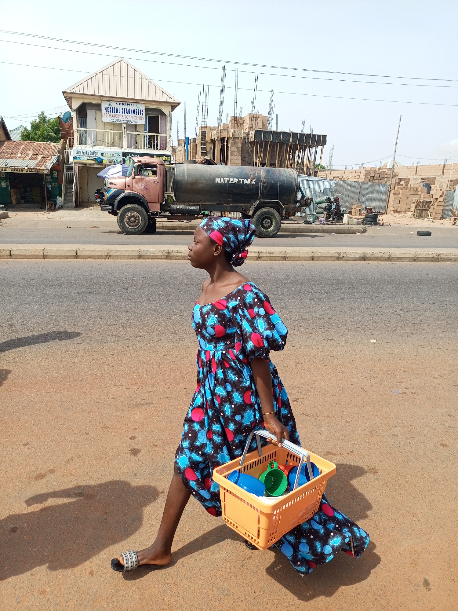 In Tempure, Nigeria, a young woman walks along a paved road, carrying an orange plastic basket filled with blue and green plastic cups. She is wearing a vibrant, patterned dress and a matching headscarf. A large, dark-colored water tank truck is parked on the side of the road behind her. In the background, buildings are visible, including one with a sign that reads "SPRING MEDICAL DIAGNOSTIC LABORATORY & SCAN CENTRE" and another under construction with scaffolding. The ground is dusty and the sky is clear.