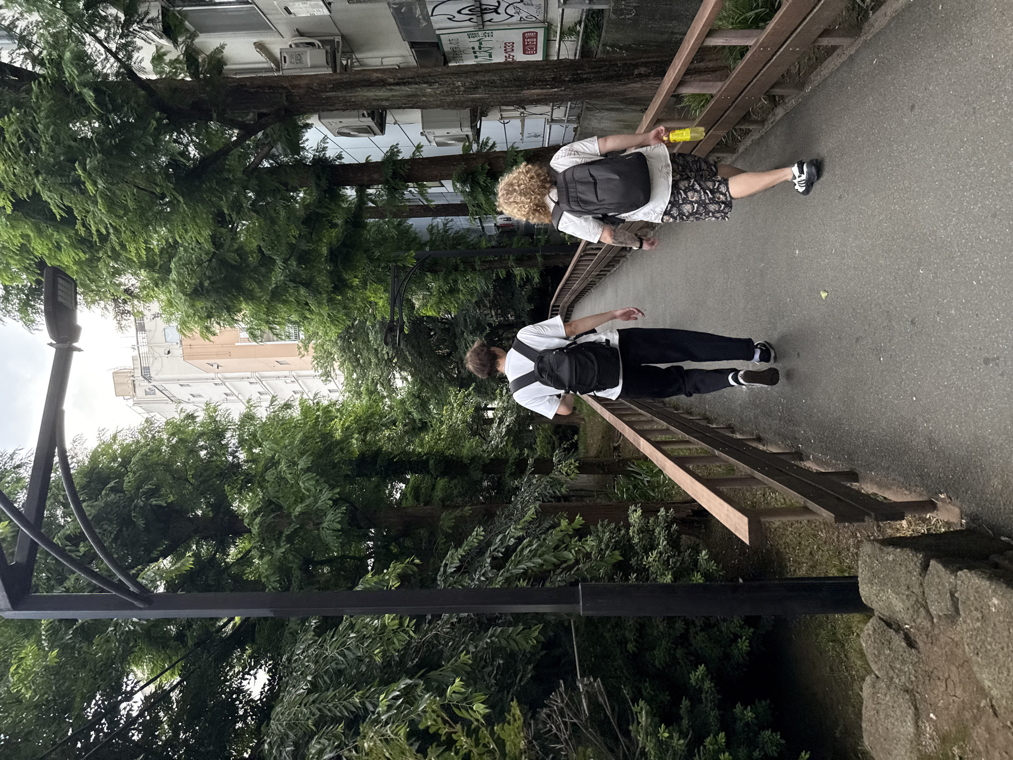 Two individuals walk away from the camera on a paved pathway bordered by a wooden railing. The pathway is flanked by lush green trees on the left and buildings on the right. The person on the left wears a white t-shirt, black pants, and a black backpack. The person on the right has curly blonde hair, wears a white t-shirt with a graphic print, patterned shorts, and a black backpack. They are also holding a yellow bottle. The environment suggests an urban setting, likely Shinjuku, Japan, with a mix of nature and city architecture. The sky appears overcast.