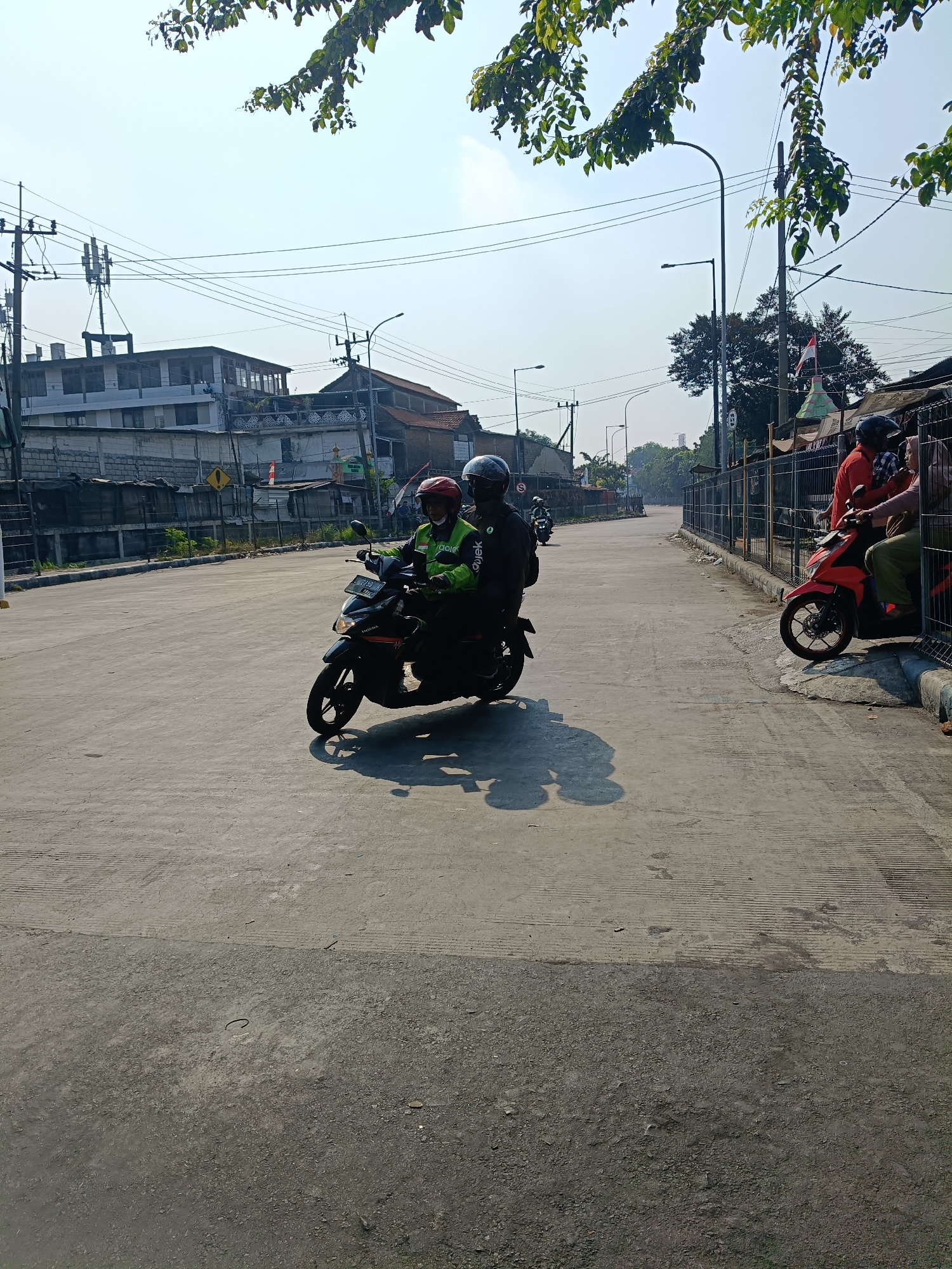 The image depicts a street scene in Kedungrejo, Indonesia, under a clear, bright sky with leafy branches of trees visible in the upper portion. In the foreground, a concrete road with distinct parallel grooves is prominent. On the road, a black motorcycle with two occupants is moving from left to right. The rider wears a red helmet and a green jacket with "Gojek" branding. The passenger wears a blue helmet. Their shadow is cast on the road.
To the right of the road, a metal fence encloses a bordered area with a sidewalk. Several people are standing near the fence. A red motorcycle is parked on the sidewalk. Two individuals, one wearing an orange shirt and the other a tan hijab and green pants, are interacting near the red motorcycle.
In the background, various buildings are visible. On the left, there are multi-story buildings with white facades and brick walls, along with electrical poles and antennas. Further down the street, more buildings and trees line the road. Streetlights are also present along the street. The overall environment suggests an urban or semi-urban setting.