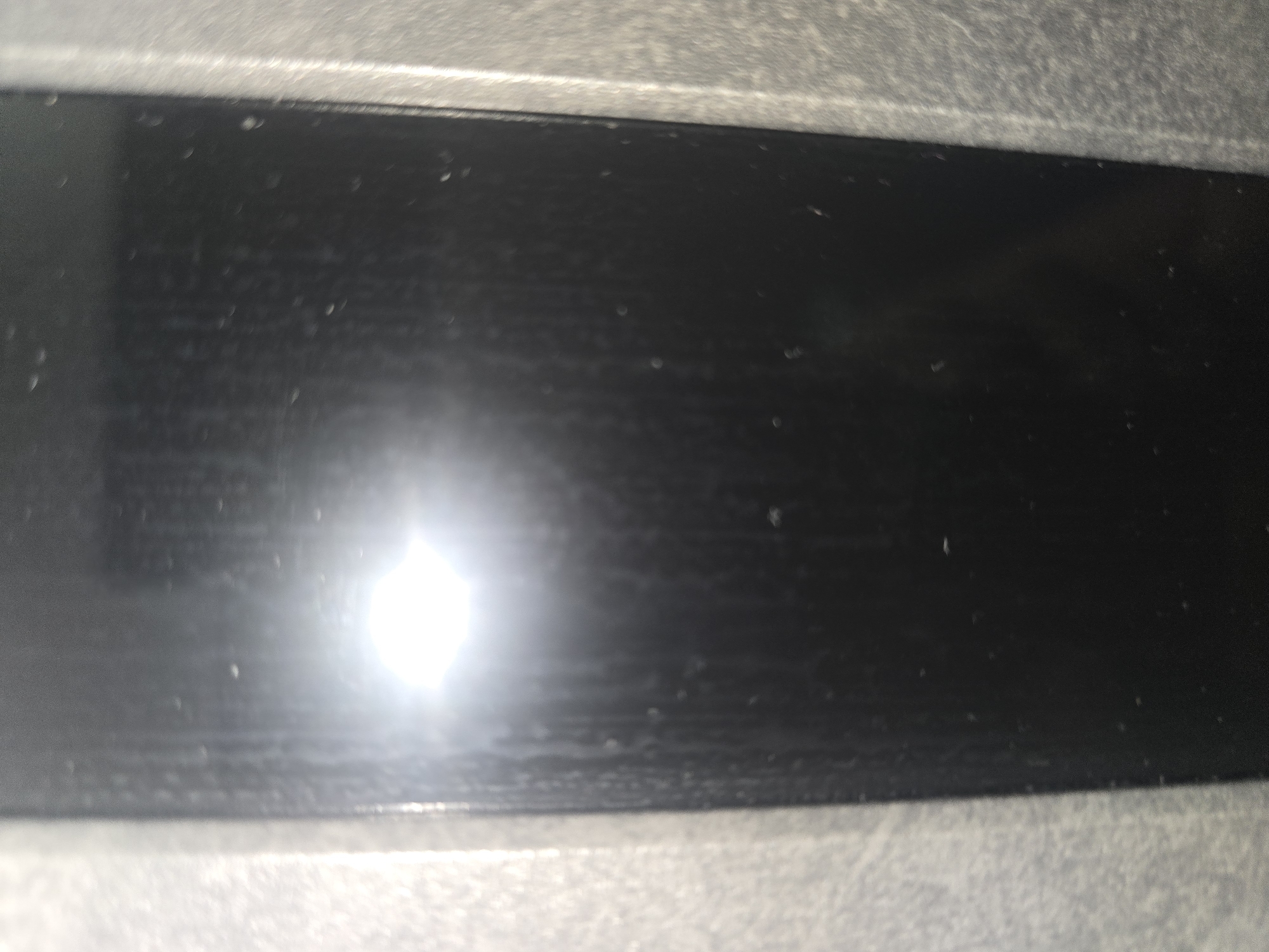 The image is a close-up, vertically oriented shot of a dark, flat surface, likely a screen or a polished material, with vertical striations visible. There are numerous small, bright specks scattered across the surface, which appear to be dust or light reflections. A large, bright, out-of-focus light source is present in the upper left quadrant of the image, creating a significant glare. The right and left edges of the image show a textured, lighter-colored material that frames the dark central surface. The scene appears to be indoors, in a dimly lit environment given the stark contrast between the bright light source and the dark surface. There is no discernible context to place this image within the city of Ulan-Ude, Russia.