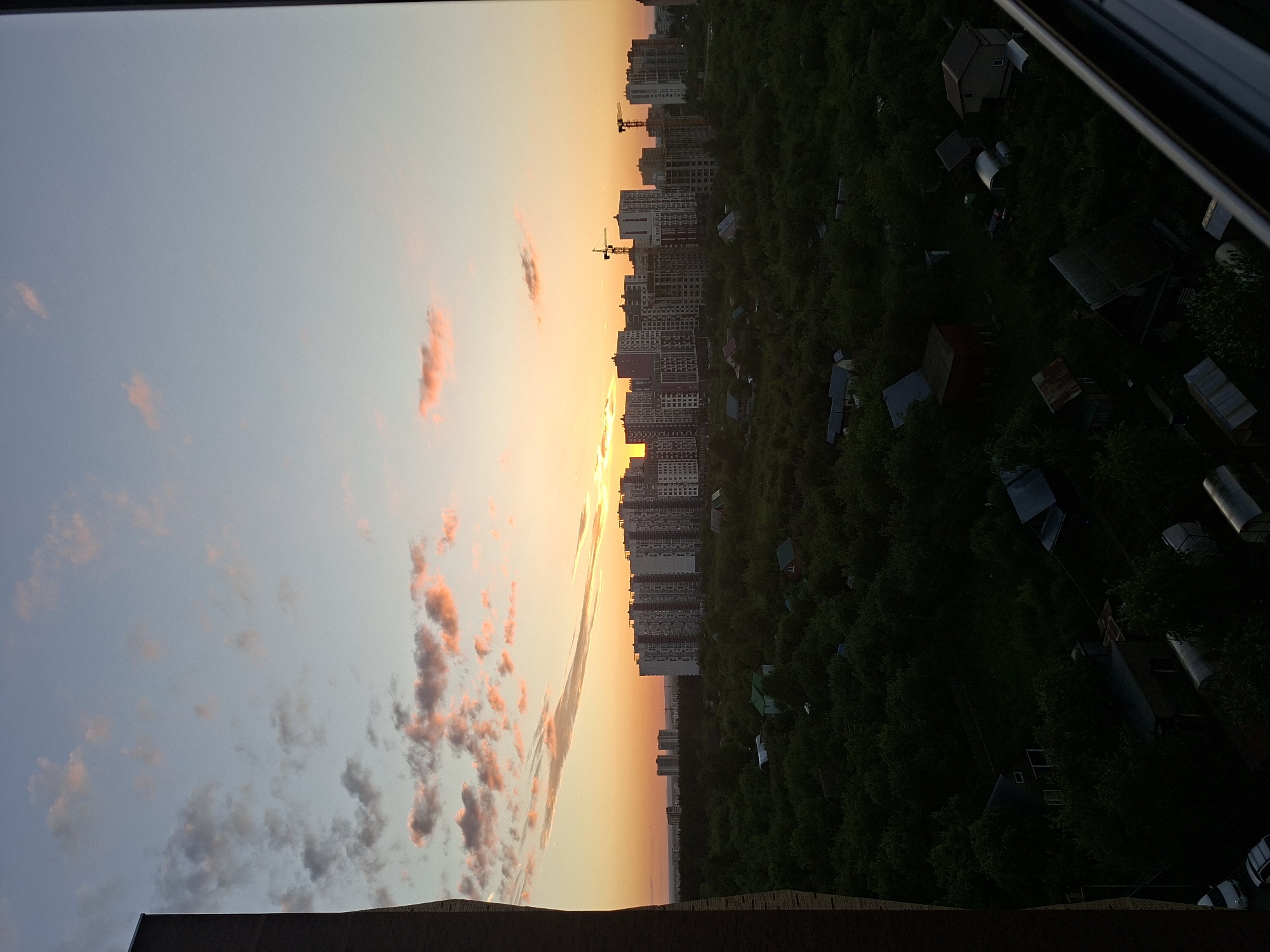 The image captures a sunset over a suburban area on the outskirts of Izhevsk, Russia. In the foreground, the dark, angular structure of a building, possibly an apartment complex, partially obscures the left side of the frame. Beyond this, a dense canopy of green trees stretches across the landscape, interspersed with numerous small, low-lying structures that appear to be dachas or garden houses. These structures are varied in design, with some featuring red or brown roofs, while others have a more rustic appearance.
In the middle ground, a cluster of multi-story apartment buildings forms a distinct urban element, silhouetted against the brightly lit sky. Several construction cranes are visible among these buildings, indicating ongoing development. The sky itself is a dramatic display of sunset colors, with a gradient of orange, yellow, and pink hues illuminating the clouds. The sun is low on the horizon, positioned behind the urban development. The overall impression is one of a transition between urban and rural, with the natural landscape giving way to modern construction under a vibrant evening sky. No people are discernible in the image.