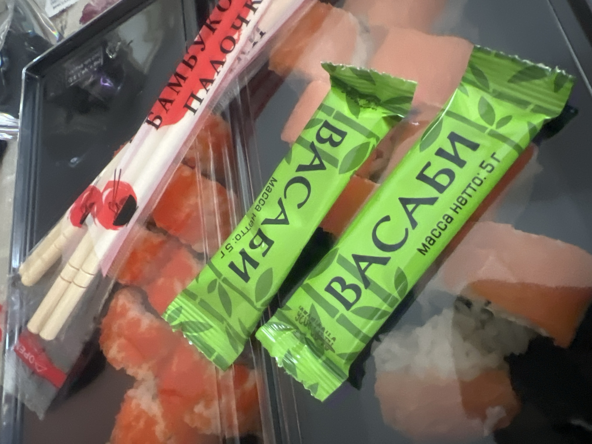 In a top-down, slightly angled medium shot, a black plastic takeout container with a clear plastic lid holds sushi rolls. Two pairs of wooden chopsticks are wrapped in clear plastic with a red and white printed design showing the word "OPEN" in red on a red tab, and a stylized image of a bowl of sushi. Two individual packets of wasabi, bright green with a repeating bamboo leaf pattern and black text in Russian, are layered on top of the sushi. The top packet's text reads "ВАСАБИ" (Wasabi) and "Масса нетто: 5 г" (Net weight: 5 g). The bottom packet also has "ВАСАБИ" visible, with the net weight likely also being 5g. The sushi rolls are visible through the plastic wrap, appearing to be salmon and avocado rolls with rice. The background is blurred and out of focus, but appears to be a flat, indoor surface. The overall lighting is bright, suggesting an indoor setting.