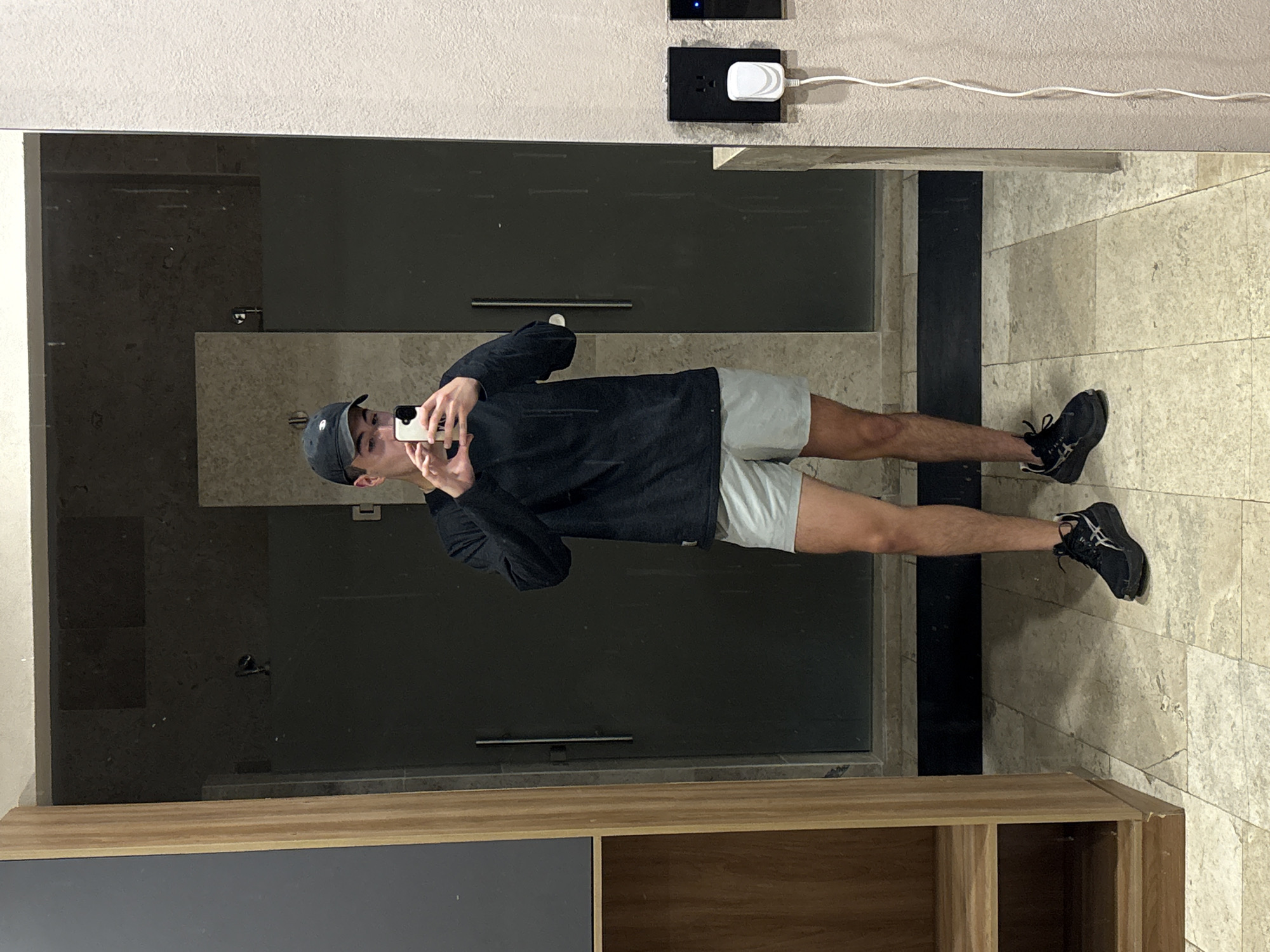A person is taking a mirror selfie in what appears to be a bathroom. The person is wearing a black cap, black long-sleeved shirt, light-colored shorts, and black athletic shoes. They are holding a cell phone up to the mirror to take the picture. The bathroom has a shower with a glass door behind the person. The wall behind the mirror has electrical outlets. The floor is tiled. A partially visible wooden and dark gray cabinet is below the mirror. The location, San Bernardino Tlaxcalancingo, Mexico, cannot be verified from the image.