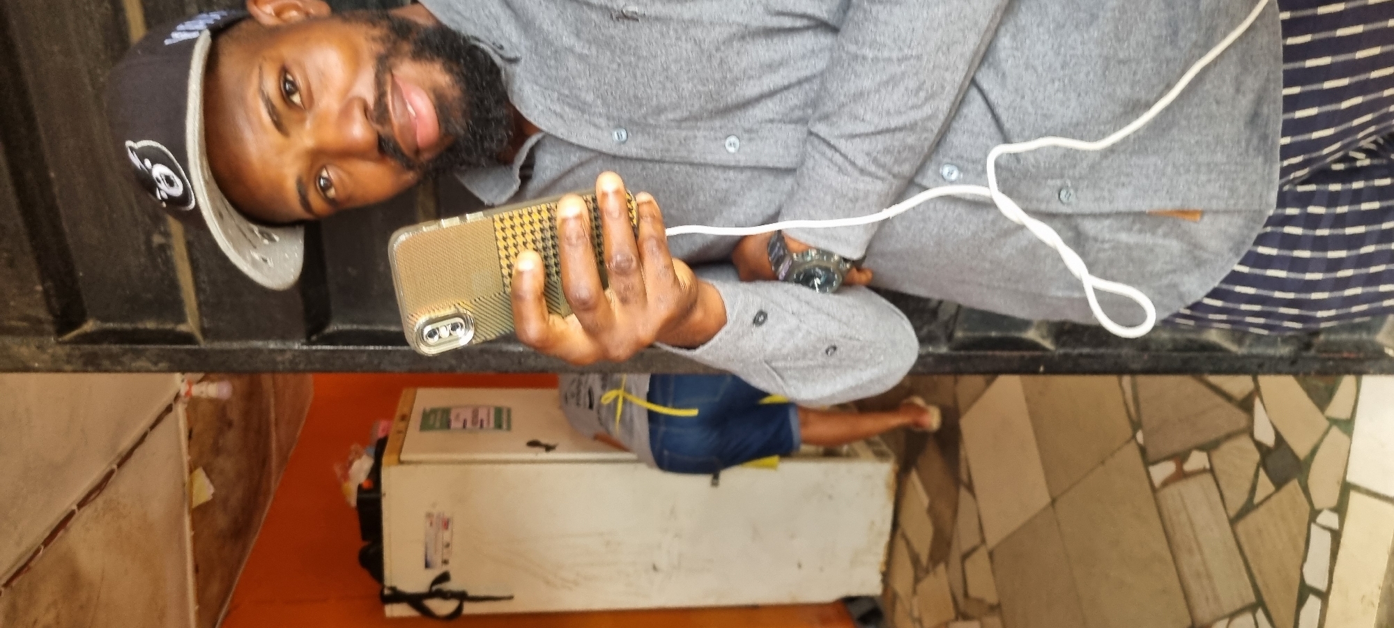 The image depicts a medium shot of a Black man wearing a grey collared shirt, a black baseball cap with a white logo, and striped pants. He is holding a smartphone in his right hand and appears to be taking a selfie or recording a video. A white cable, possibly headphones or a charger, is visible trailing from the phone. The background is partially obscured by a dark doorway. To the left, a white refrigerator is visible, and behind it, another person wearing denim shorts and a yellow apron is bending over. The floor is tiled with a mosaic pattern. The lighting suggests an indoor or semi-outdoor setting, possibly in a residential or commercial space within Lagos, Nigeria. The overall environment appears utilitarian.