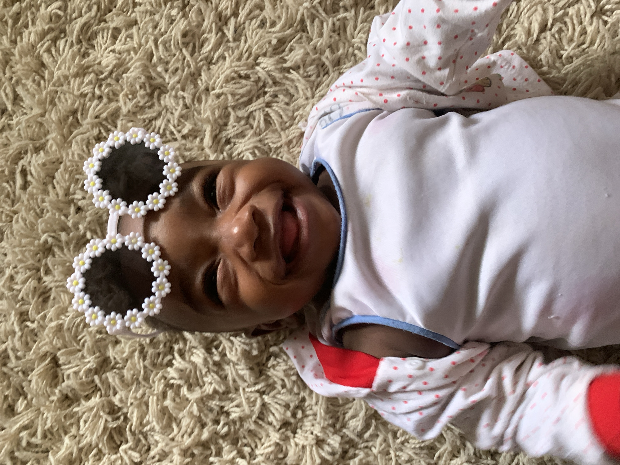A baby lies on a beige shag carpet. The baby wears a white onesie with red polka dots on the sleeves and a red stripe on the right side, and flower-shaped sunglasses. The baby is smiling and looking at the camera. The location appears to be indoors, potentially a home. It's impossible to confirm the location in Barakin Akawo, Nigeria, based solely on the image.
