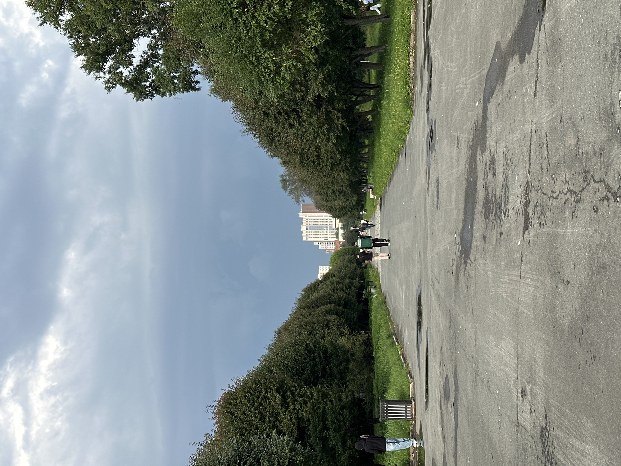 This image shows a wide, paved pathway in a park, bordered on both sides by rows of mature trees. The pathway appears wet and has puddles, suggesting recent rain. In the distance, several modern, multi-story buildings are visible, indicating an urban setting. A few people are walking on the pathway. On the left, one person in a black hooded jacket and jeans is standing near a trash can. Further down the path, two individuals, one in a green shirt and the other in black, are walking away from the camera. The sky is overcast with dark, heavy clouds. This scene is likely from the Yekaterinburg Central Park of Culture and Recreation named after V.V. Mayakovsky, a well-known park in Yekaterinburg, Russia, due to the combination of a park setting with tall buildings in the background.