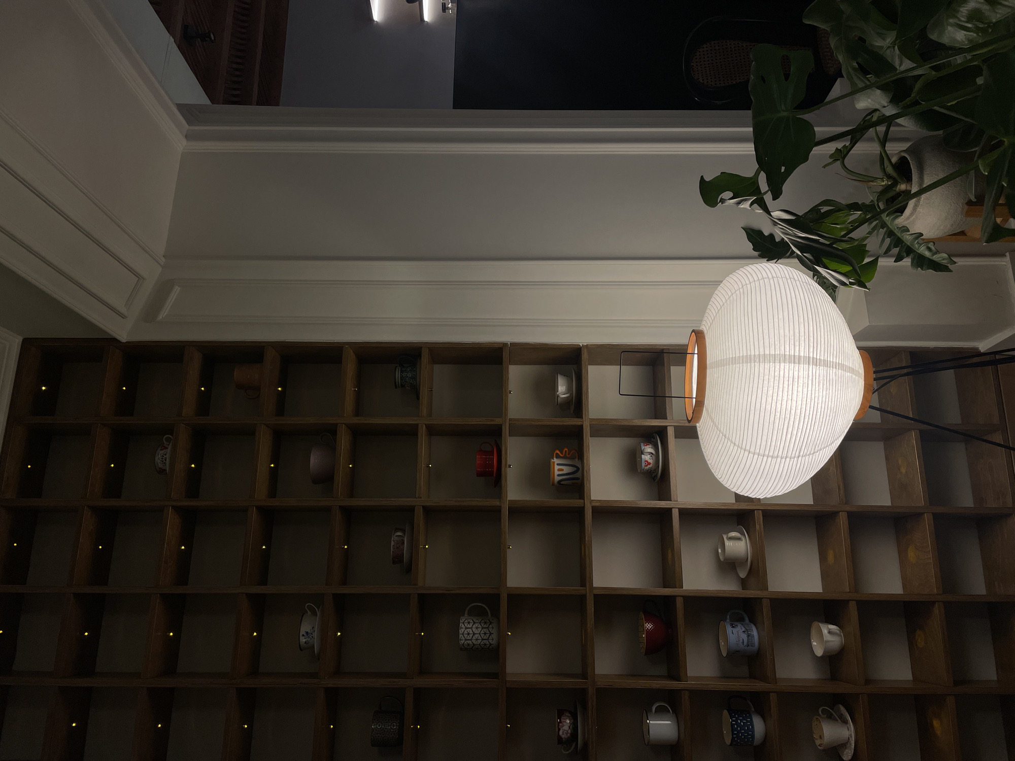 The photograph depicts an interior space in Yerevan, Armenia. A large, custom wooden shelving unit fills the lower half of the frame, displaying various coffee cups or mugs in different styles, and small golden light elements. Above, a white wall with decorative molding is visible. A large, round paper lantern dominates the right side of the image, partially obscuring the shelving. Green plants with large leaves are positioned above the lantern, suggesting a decorative arrangement. The lighting in the scene is dim, highlighting the contrast between the lantern and the darker shelves.