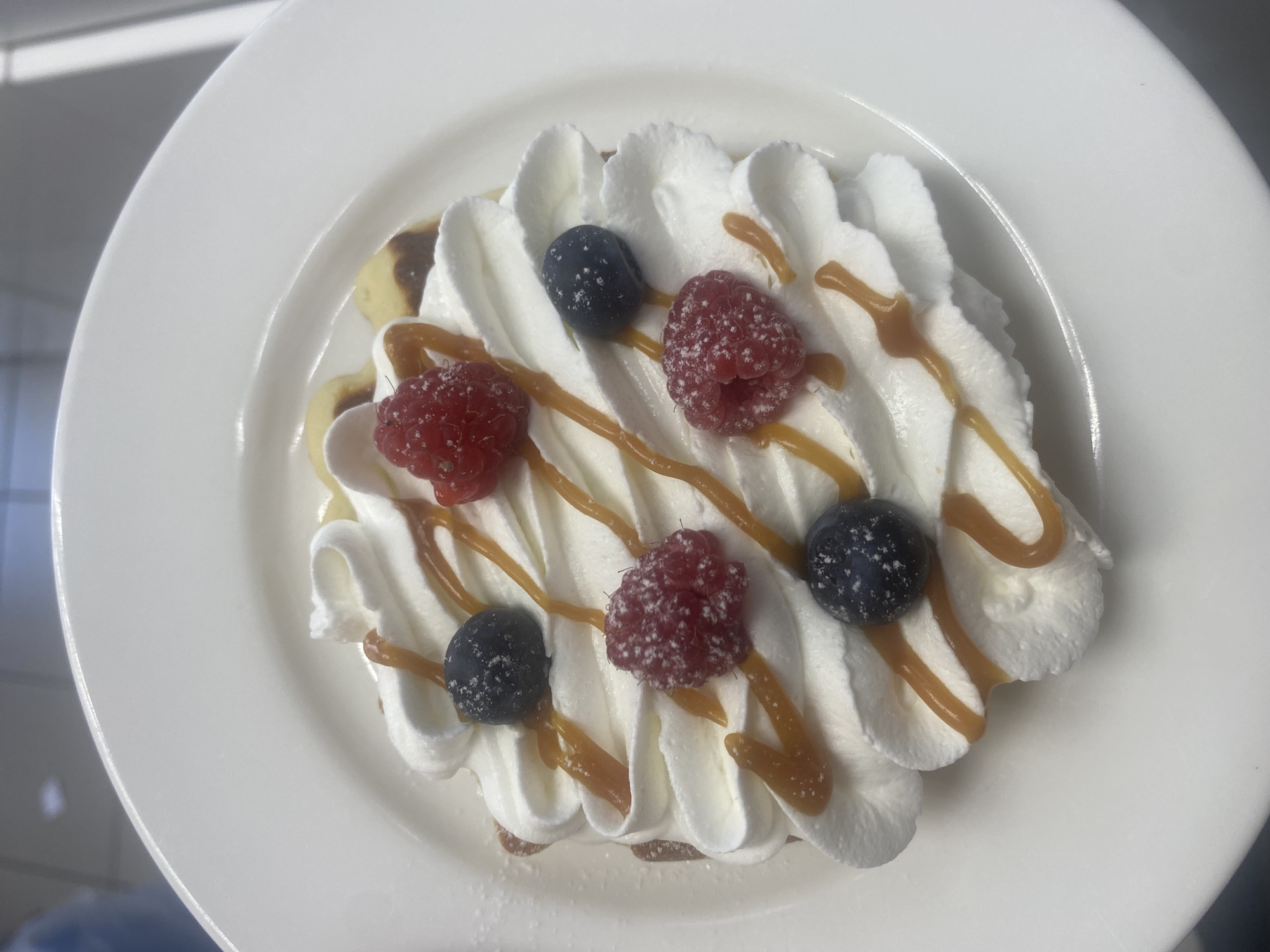 A top-down view captures a dessert served on a white plate. The dessert features several small pancakes topped with a generous swirl of whipped cream. Drizzled over the whipped cream are thin lines of caramel sauce. Scattered atop the cream are fresh raspberries and blueberries, some dusted with what appears to be powdered sugar. The overall presentation suggests a sweet and indulgent treat. The setting appears to be indoors, possibly a restaurant or kitchen, given the clean white plate and the indistinct background. The lighting is bright and even, indicating daytime or well-lit interior. No people are visible in the frame, and no text is present. The focus is solely on the appetizing dessert. The dessert is a single serving, presented neatly on a round white plate. The whipped cream is piped in decorative peaks, creating a visually appealing texture. The caramel drizzle adds a golden contrast to the white cream and red and blue berries.