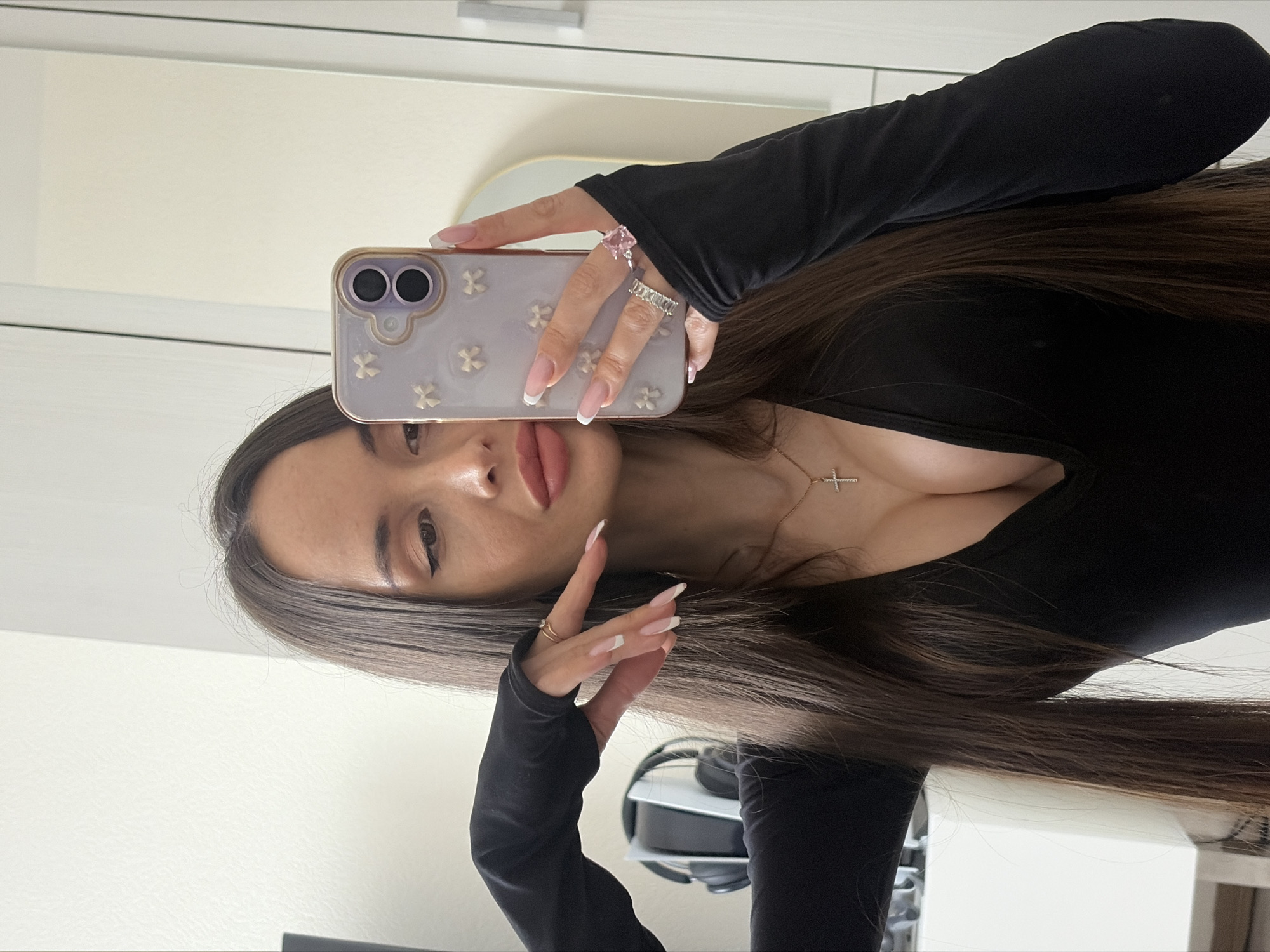 A brunette woman is taking a selfie with her phone. The phone has a light purple case with a floral pattern and a gold-colored frame. She is wearing a black long-sleeved shirt with thumb holes. She has manicured nails with a white tip. She is wearing multiple rings on her fingers. She is wearing a gold necklace with a cross pendant. In the background, there is a white cabinet with silver handles, a light yellow curved object, and a white and black gaming headset on a white stand. The background is a white wall. The location is not discernible from the image.