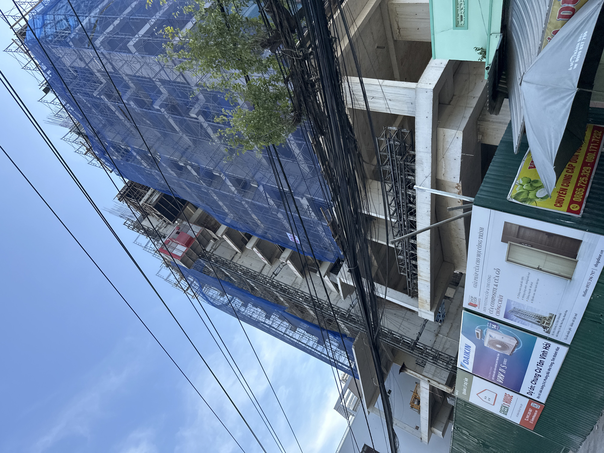 This is an outdoor, eye-level, wide-angle shot of a building under construction in Bac Nha Trang Ward, Vietnam. The structure is a multi-story concrete building, partially covered by blue netting and scaffolding. A red elevator shaft is visible on the side of the building. Power lines crisscross the sky in the foreground. At the base of the construction site, a green fence displays several signs. One sign features the "GREEN'S HOUSE" logo and contact information. Another sign shows a "DAIKIN" air conditioning unit. A third sign advertises "SHOP DOOR" and lists features like "CHONG CHAY" (fire-resistant) for composite and wooden doors. A fourth sign, partially visible, promotes a coconut sale. The overall impression is of an active construction site in an urban environment.