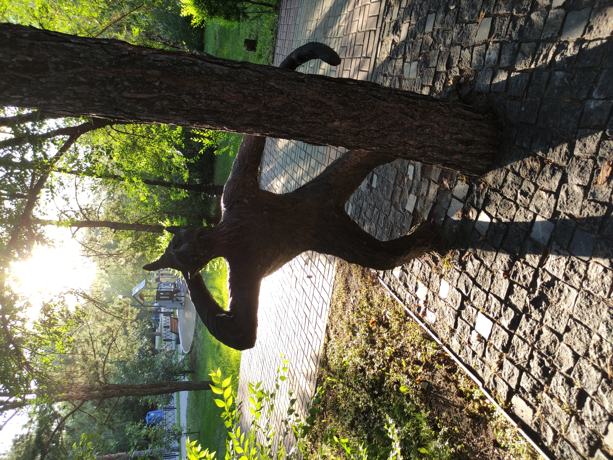An outdoor shot in Semey, Kazakhstan, captures a dark, abstract statue that resembles a humanoid figure with animalistic features, perhaps a cat or a wolf. The sculpture is positioned beside a tree, with one of its arms raised to its head. The ground around it is paved with rough, irregular stones, and a pathway of lighter, rectangular stones is visible further away. Sunlight filters through the leaves of trees, casting dappled shadows. In the background, a playground with swings and slides can be seen, along with lush green grass and more trees. A blue recycling bin is visible on the left side of the frame. The scene appears to be a park or public outdoor space.