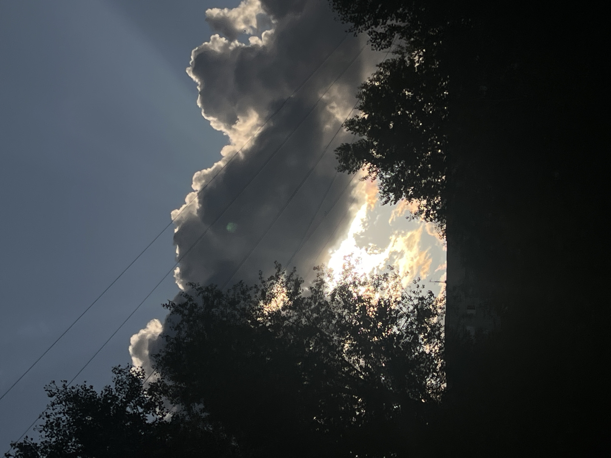 This image captures a dramatic sky scene with dramatic clouds silhouetted against a dark sky. The sun is breaking through the clouds, creating bright rays and illuminating the edges of the clouds with a golden hue. Overhead, power lines crisscross the frame, adding a man-made element to the natural spectacle. In the foreground, the dark silhouettes of trees and a building suggest a setting in an urban or suburban area. The image was taken on a clear day with a light breeze. The overall mood is one of awe and natural beauty.