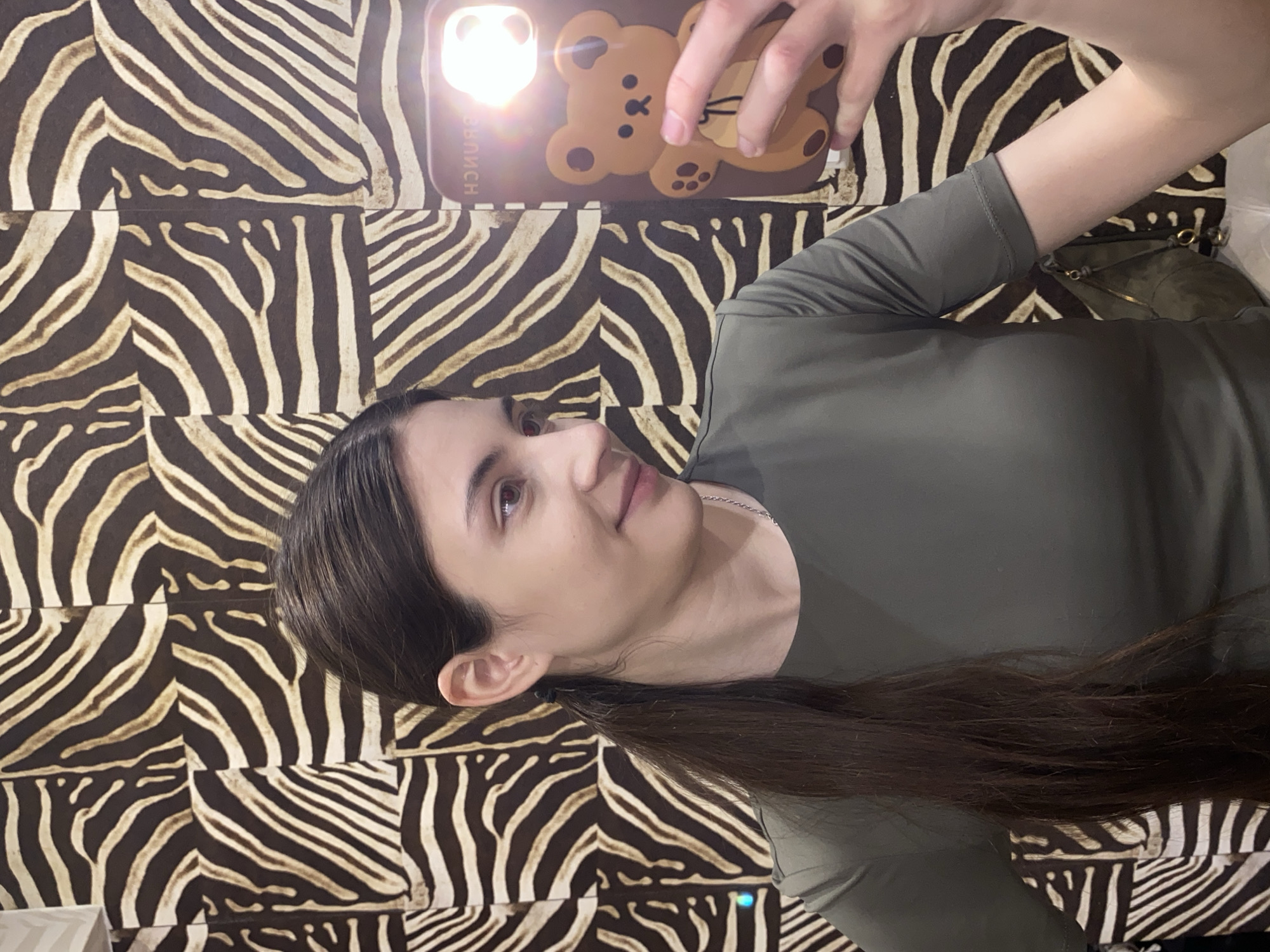 The image features a young woman taking a selfie in an indoor setting in Votkinsk, Russia. She has long, dark brown hair that falls over her shoulder, fair skin, and dark eyes, looking upwards towards the camera with a subtle, pleasant expression. She wears a dark olive green long-sleeved top and a thin silver chain necklace.
She holds a smartphone with her right hand, positioned above her head. The phone is encased in a light brown cover adorned with a cheerful cartoon bear design, complete with heart-shaped paw pads. A bright flash or light source emanates from the top left corner of the phone, illuminating her face. Due to the mirror effect of the selfie camera, the word "BRUNCH" is visible in reverse text on the phone case.
The background is a prominent zebra-print pattern of dark brown and light beige irregular stripes, suggesting a wallpaper or fabric covering on a wall. Partially visible behind her right arm is what appears to be a dark green or olive-colored handbag with gold-colored metal details. The overall image is rotated 90 degrees clockwise, indicating a casual moment captured by the woman herself.