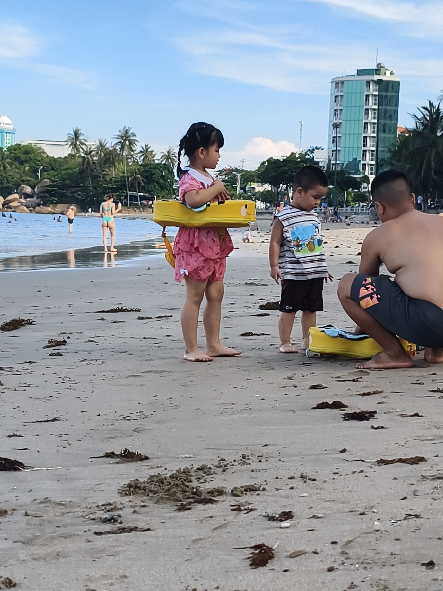 **Scene:** Beach in Bac Nha Trang Ward, Vietnam.
**People:**
A man is kneeling on the beach. Nearby, a young girl wearing a pink dress and a yellow object around her waist. A young boy stands next to her. Two women in swimsuits are in the water. Other people visible in the background.
**Objects:**
A suitcase is present on the sand. Palm trees, building, and other beach features.
**Actions:**
The man appears to be interacting with the suitcase. The children are standing on the sand.
**Environment:**
Beach setting with sand, seaweed and clear sky. The water has gentle waves.