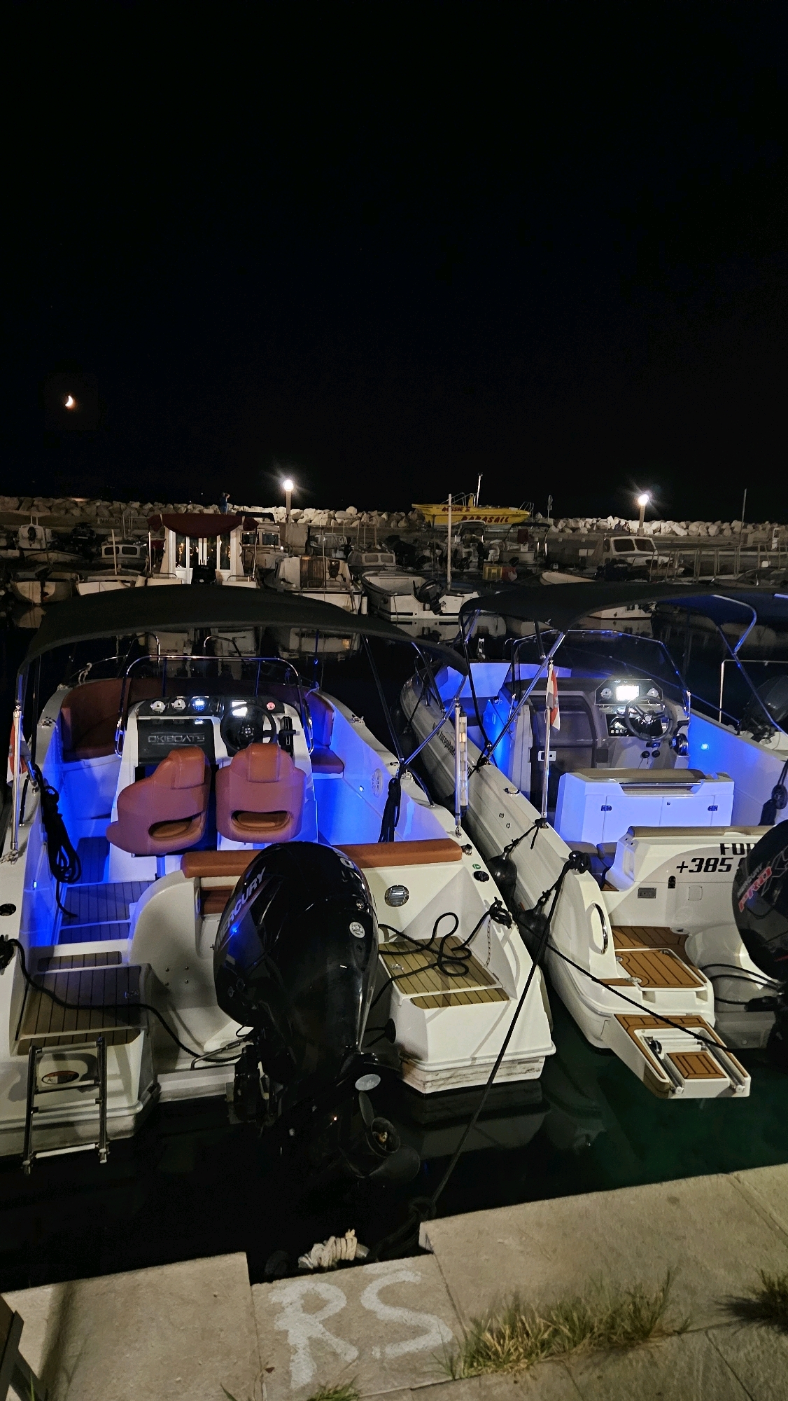 At night, several motorboats are docked at a marina in Općina Tučepi, Croatia. The boats are illuminated by artificial lights, creating a vibrant blue glow within their cabins. The scene is dark, with the exception of the boat lights, a crescent moon visible in the sky, and some distant streetlights. The boats are moored next to each other in calm water. In the foreground, the concrete edge of the dock is visible with the spray-painted initials "RS". In the background, a rocky shoreline can be seen, with more boats and some buildings.