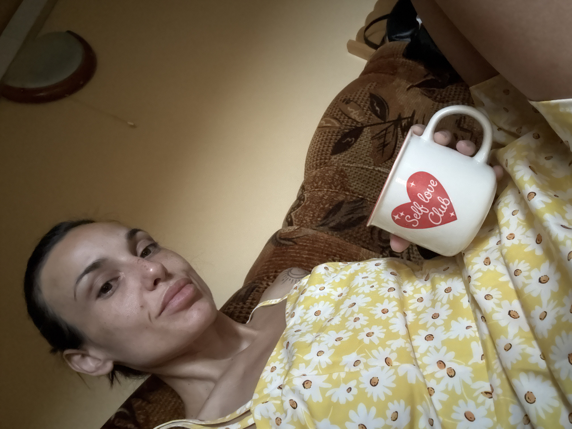 In Pleven, Bulgaria, a woman with dark hair pulled back sits on a patterned sofa, holding a white mug with a red heart and the words "Self love Club." She is wearing a yellow dress with a daisy print. The background consists of a beige wall and a portion of the sofa's armrest.