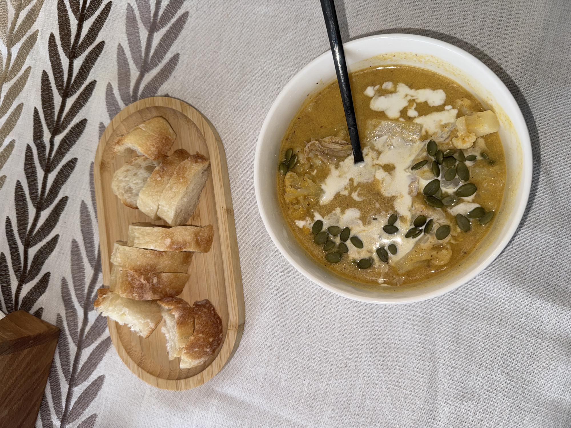 The scene depicts a meal consisting of a bowl of soup and sliced bread served on a table covered with a patterned tablecloth. The soup, in a white bowl, appears to be a creamy yellow color, possibly pumpkin or squash, with a garnish of cream and green seeds. A black spoon is in the bowl. The bread is sliced and arranged on a light-colored wooden board. The location is unknown and cannot be determined from the image. No people are visible. The objects are the soup bowl, soup, spoon, sliced bread, wooden board, and tablecloth. There are no apparent actions or interactions. The environment is an indoor dining setting.