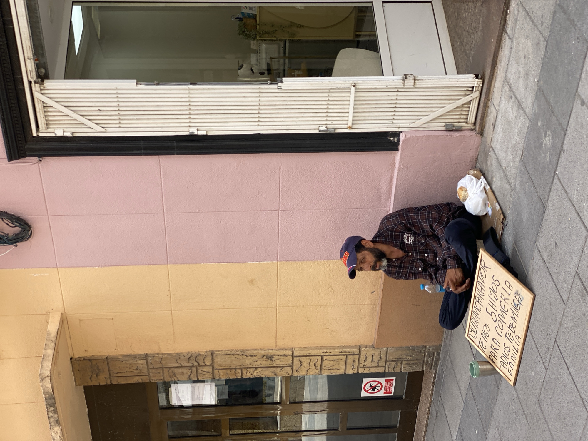 A man sits on the sidewalk in front of a building with a pastel pink and yellow facade. He wears a plaid shirt, dark pants, and a blue baseball cap. In front of him is a cardboard sign that reads, in Spanish, "AYUDAME POR FAVOR TENGO 5 HIJOS PARA COMER LA FADIO TEBEMIGA FAMILIA" which translates to "HELP ME PLEASE I HAVE 5 CHILDREN TO EAT THE FAMILY'S FADIO TEBEMIGA". Next to the sign is a metal cup, a plastic water bottle, and a plastic bag containing a piece of bread. A red and white sign with a graphic of a dog on a leash is affixed to a glass door on the left. The man is looking to his right with his legs crossed. The door to the right appears to be a storefront with a metal security gate. The lighting suggests it is daytime. The scene is captured from a medium-low angle.