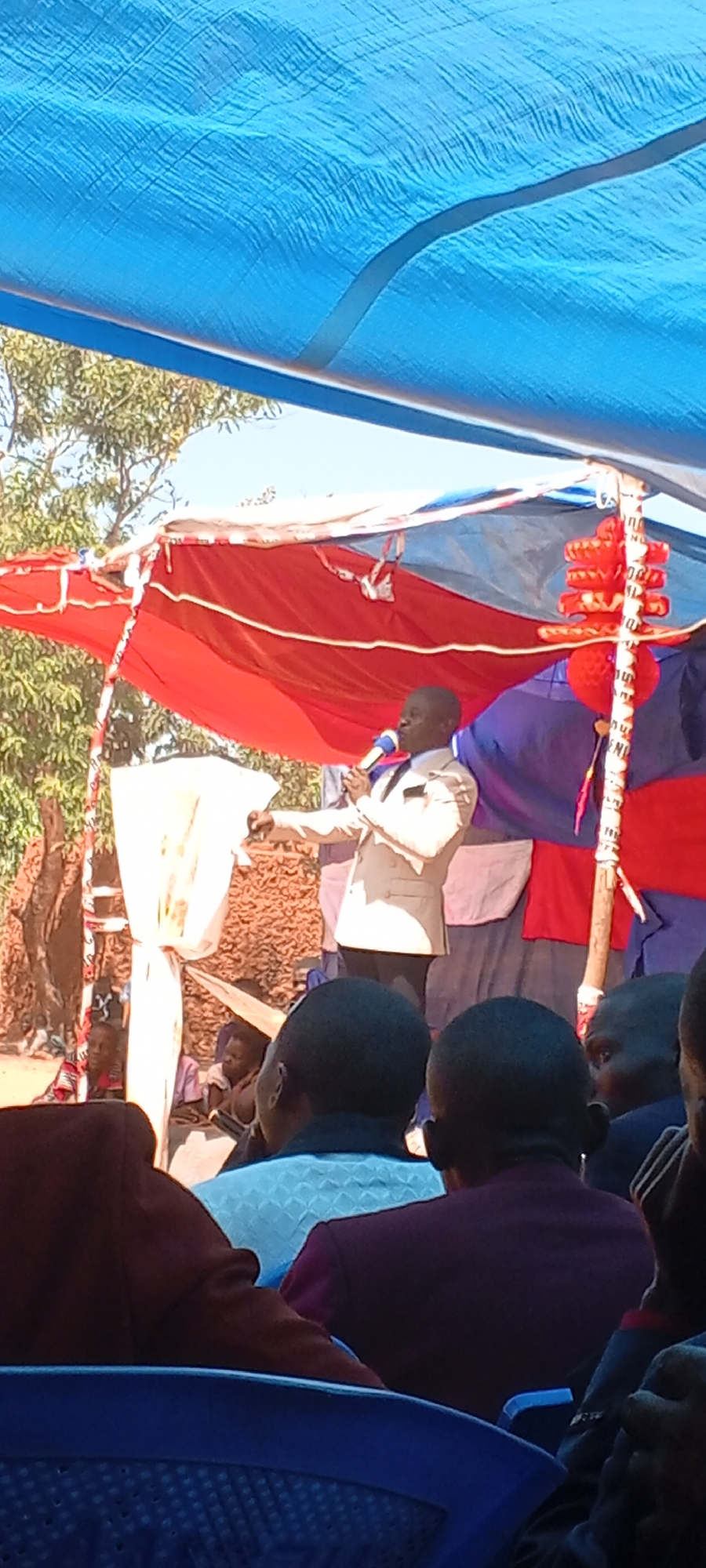 The image depicts a speaker addressing a crowd under a makeshift canopy, possibly in an outdoor setting in Kalemie, Democratic Republic of the Congo. The canopy is made of blue and red tarps supported by wooden posts. The speaker, wearing a light-colored suit, is holding a microphone and gesturing with his right hand. The audience, seated in blue plastic chairs, appears to be attentive. The environment includes trees and other vegetation in the background, suggesting a rural or semi-rural location. Additional people are sitting on the ground behind the speaker. Red decorative tassels are visible near one of the support poles. The overall impression is that of a public gathering or event.