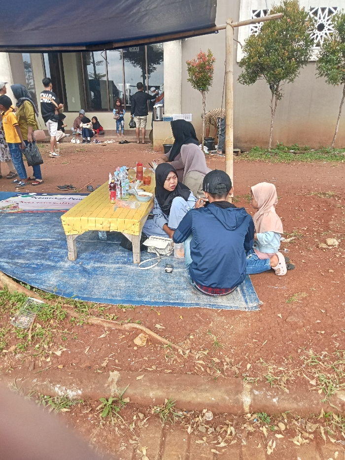 This outdoor scene in Singaparna, Indonesia, depicts a casual gathering of people under a blue tarp awning, likely enjoying a meal or snacks during the daytime. The ground is unpaved, reddish soil, with some grass and exposed tree roots.
In the foreground, a group is seated on a blue tarp spread on the ground. A low, yellow-covered table holds various food items in bowls, plastic containers, and bottled drinks (including water and sauce). A woman in a black hijab and light blue top is seated on the left, looking towards the right and engaged in conversation. To her right, a man in a blue jacket and black cap is seated with his back to the camera. Next to him, a small child in a pink hijab and light blue top is also seated on the ground, facing the man. Another woman in a light brown hijab is partially visible behind the woman in black, also seated. A white handbag and a small toy car are visible on the blue tarp near the seated individuals.
Further back, more people are gathered. Several individuals stand near a building with large windows reflecting the sky and surrounding trees. One man in a black t-shirt and light shorts is prominent among the standing group. Others are scattered, some seated on the ground or low structures. The building itself is light-colored with decorative cut-outs on a section of its wall, and small trees are planted around it. The blue tarp awning under which the foreground group is seated is supported by rough wooden or bamboo poles.
The overall atmosphere appears relaxed and social, with people interacting. The clear lighting indicates it's daytime, and the activities suggest fair weather. Faint text is visible on the blue tarp in the foreground, reading "KEMERIAH KAN PENG...", possibly part of a banner or mat indicating an event or celebration.