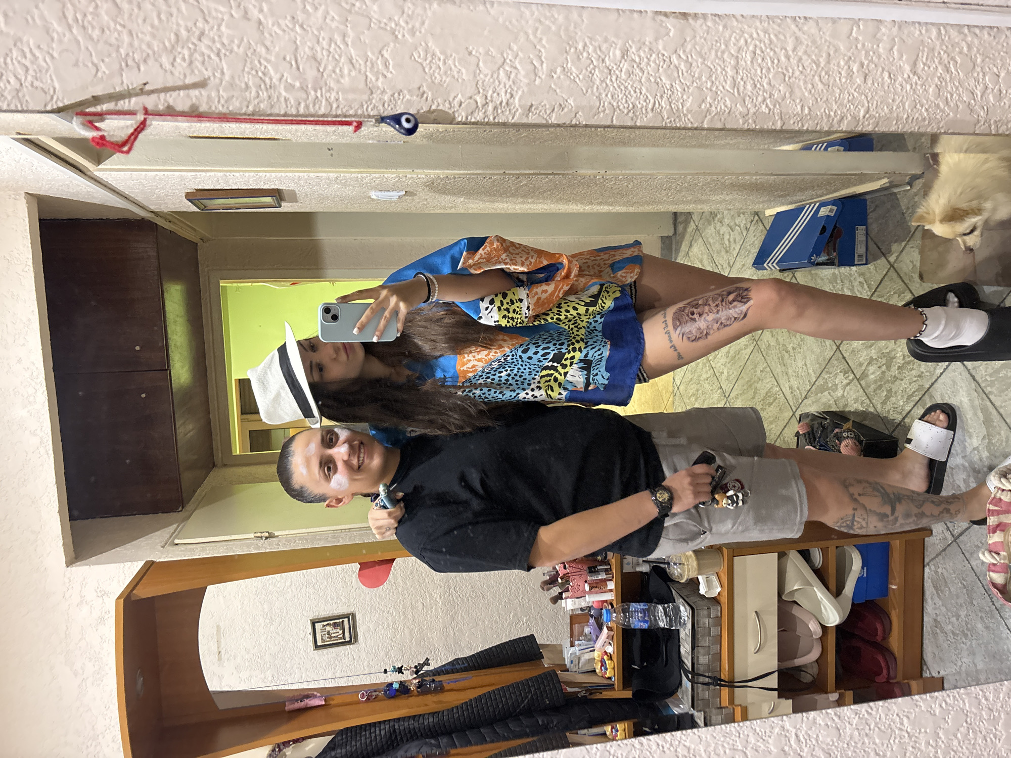 Two individuals are taking a mirror selfie in what appears to be an interior living space. The person on the left has their arm around the shoulder of the person on the right. The person on the left is wearing a black t-shirt, grey shorts, and white slides, with a watch on their left wrist. The person on the right is wearing a white hat, a blue and orange patterned kimono-style top, and black shorts. They are holding a smartphone, taking the selfie. There are various items of furniture and personal belongings visible in the background, including a mirror-backed cabinet, shelves with toiletries and shoes, a small dog, and a shoe box. The walls are textured. The presence of an Adidas shoe box and a general aesthetic suggests a casual, residential setting, possibly an apartment or house. No specific identifying features of Sofia, Bulgaria are visible within the frame.