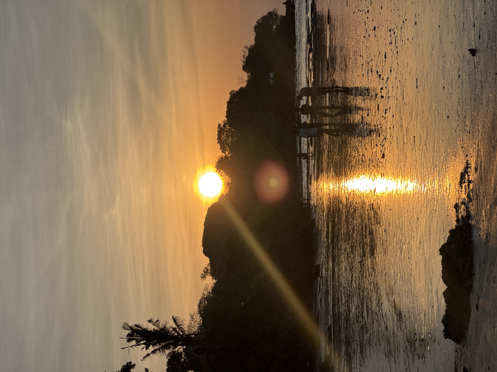 The image depicts a sunset scene at a beach in Kuta Selatan, Indonesia. The sun, a bright orb, is setting on the horizon, casting a golden glow across the sky and water. Silhouettes of trees and foliage line the elevated coastline in the background. In the foreground, several individuals are visible on the wet sand and shallow water, appearing to be walking and interacting. The wet sand reflects the vibrant colors of the sunset, creating a shimmering effect. Rocks and debris are scattered across the beach.