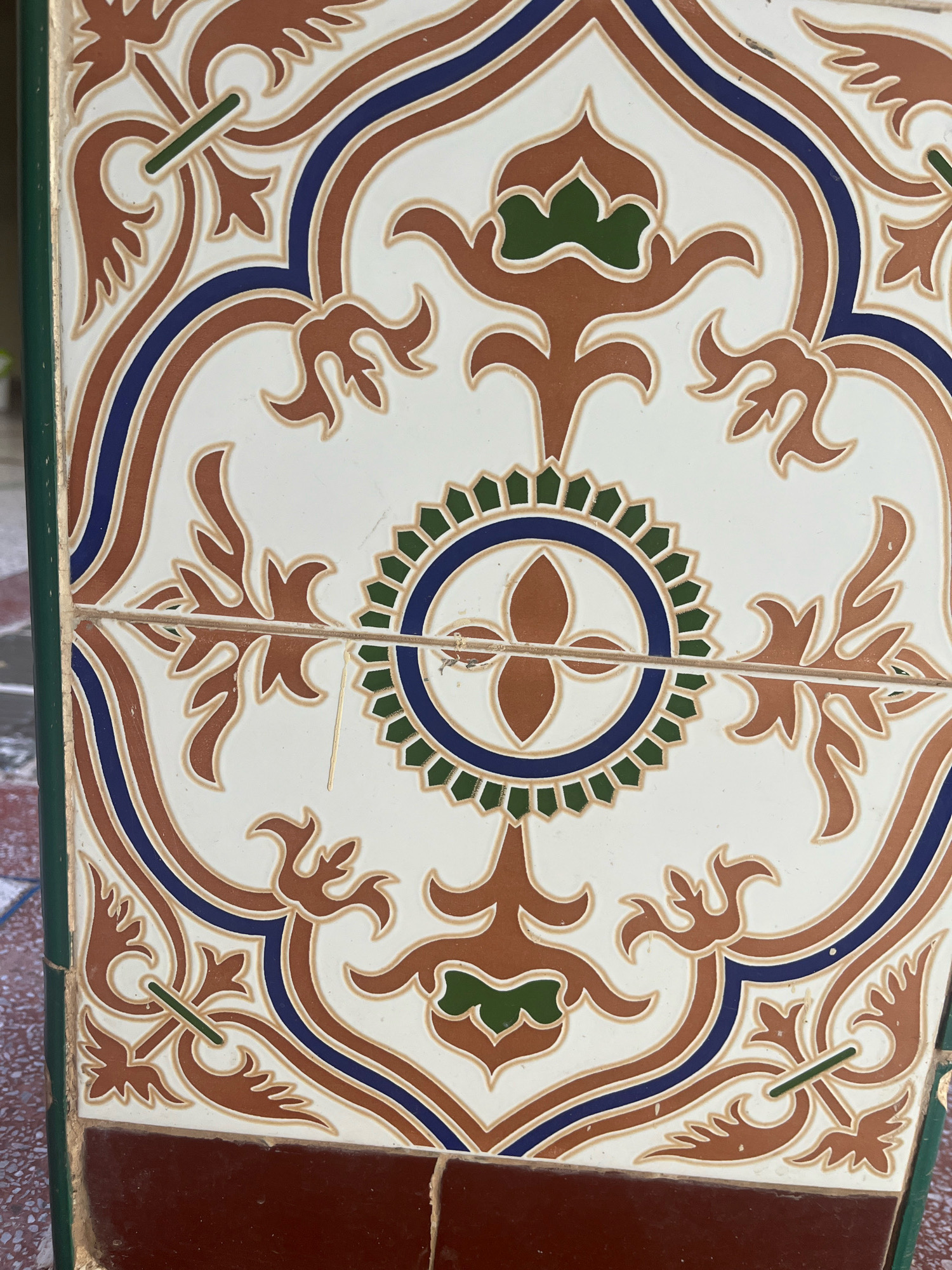 The image shows a close-up of a decorative tile in Azilal, Morocco. The tile is white with intricate patterns in brown, blue, and green. The design features floral motifs and geometric shapes, characteristic of traditional Moroccan tilework, often found in homes, riads, or public spaces. The tile is part of a larger installation, with visible grout lines indicating multiple tiles are present. The surrounding environment is not clearly visible, but the presence of tiles suggests an architectural setting. The image is sharp and well-lit, allowing for clear observation of the tile's details.