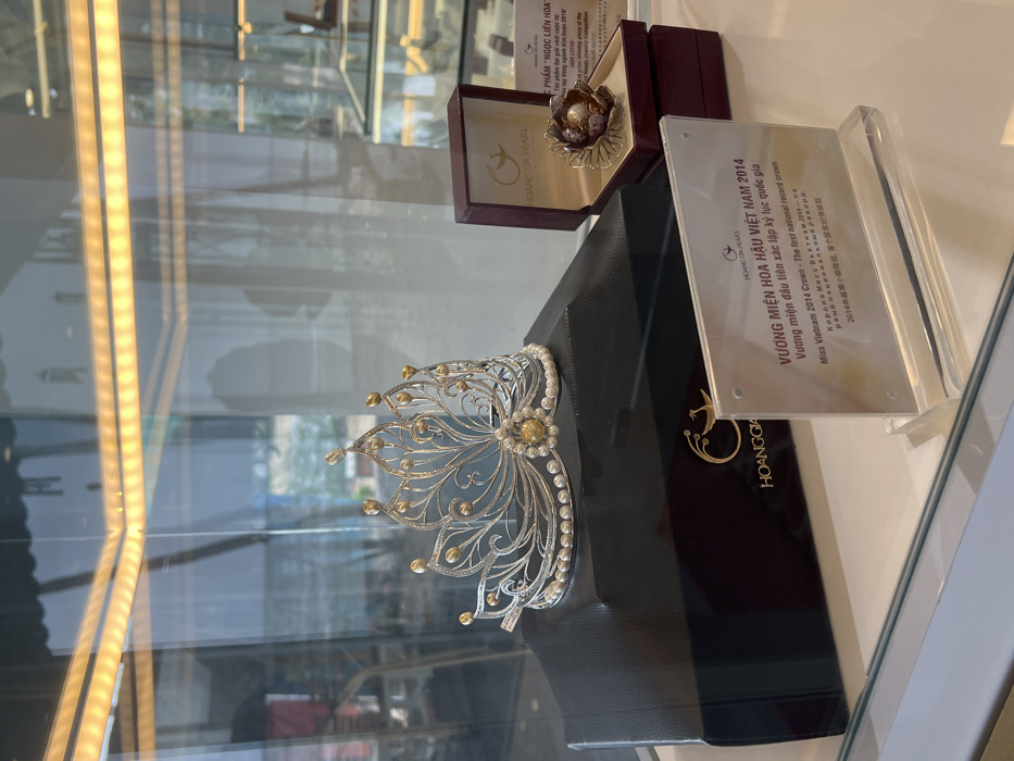 A display case contains pearl jewelry. A pearl-adorned crown sits atop a black display block next to a plaque labeled "HOANG GIA PEARL" and "VƯƠNG MIỆN HOA HẬU VIỆT NAM 2014" indicating it is the Miss Vietnam 2014 Crown. Behind it, a boxed piece of jewelry labeled "NGỌC LIÊN HOA" is visible. The case's glass reflects the surrounding environment and overhead lighting.