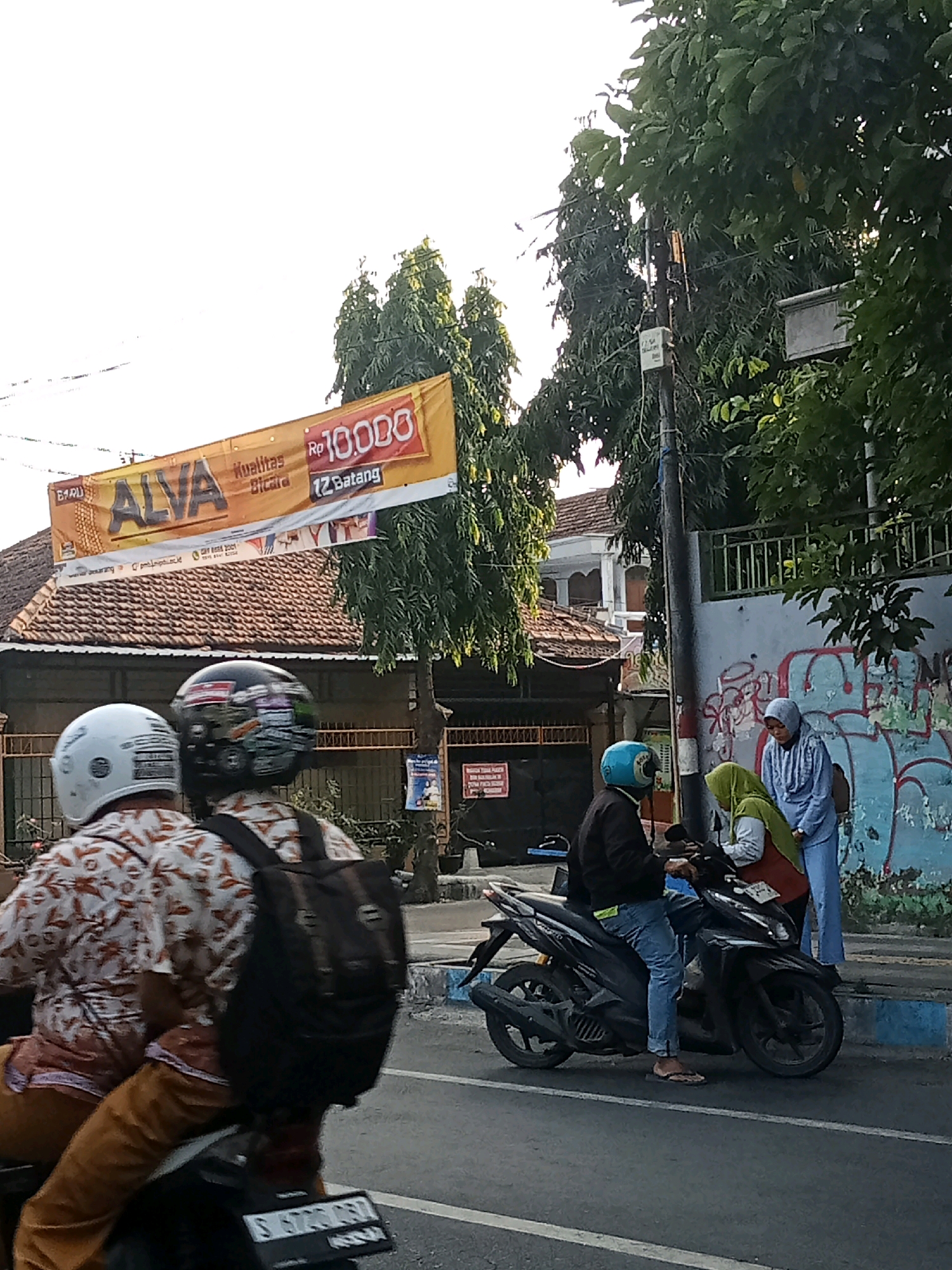 A yellow banner with red lettering promoting "ALVA" and "Rp 10,000" is suspended above a street in Jombang, Indonesia, in front of a building with a tiled roof. Several people are present in the scene. Two individuals on a motorcycle are seen from behind, wearing patterned shirts and helmets. Another person is seated on a motorcycle, wearing a dark jacket and blue jeans, while two women, one in a green headscarf and the other in a blue headscarf, stand near the motorcycle, interacting with the seated individual. The background shows trees, a utility pole with signage, and a wall with graffiti. The overall scene appears to be a street environment with commercial and residential elements.
