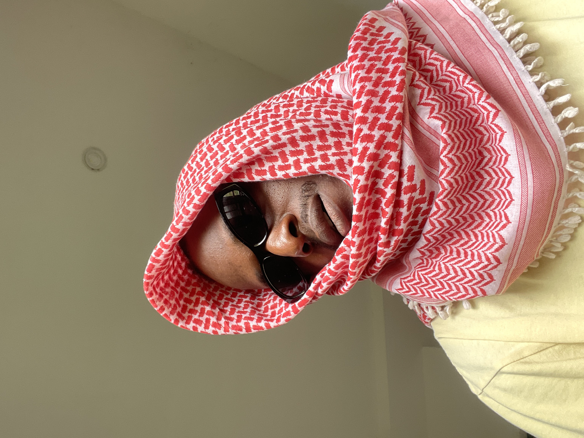 A medium shot captures a person from the chest up, looking down and slightly to the left. They are wearing black sunglasses, a red and white patterned keffiyeh draped over their head and shoulders, and a pale yellow t-shirt. The background is a plain, light-colored wall, suggesting an indoor setting. The lighting appears to be natural, coming from above and to the right, casting subtle shadows on the subject's face and the keffiyeh. The image has a slightly grainy texture. There is no visible indication of the location being in Abuja, Nigeria.