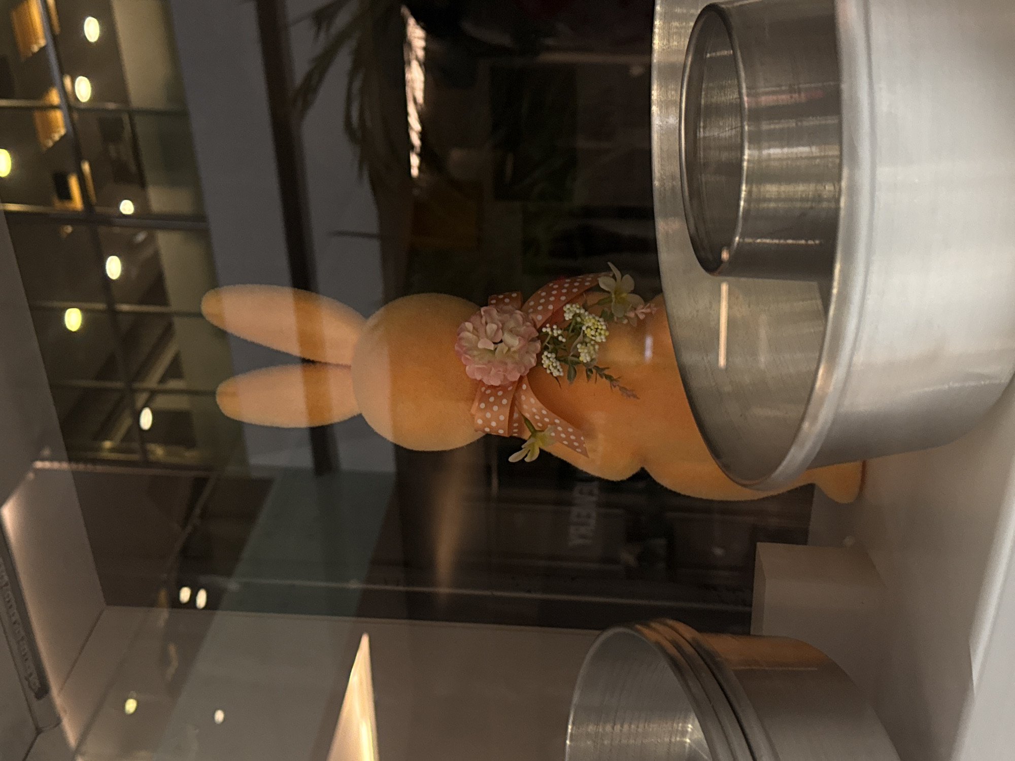 The image shows a stuffed bunny with flowers attached to its neck, positioned behind a metal object that appears to be a baking pan. The view is through glass, with blurred reflections of lights and architecture, suggesting the bunny is in a display case or window. The scene is difficult to pinpoint in Puebla without further context or visual landmarks clearly visible in the reflections. More information about the location is required to specify its location within the city.