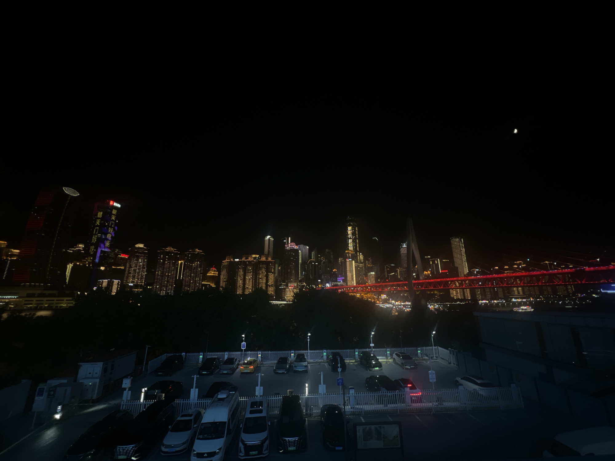 The image shows a nighttime view of the Jiangbeicheng Subdistrict in China, featuring a city skyline with illuminated skyscrapers and a bridge. In the foreground, there is a parking lot with several cars parked. The overall scene depicts an urban landscape at night, with the lights of buildings and vehicles contributing to the illumination.