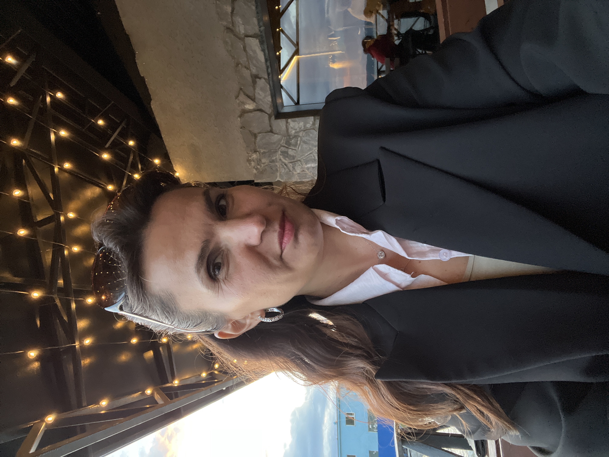 A woman is taking a selfie outdoors at dusk. She is wearing a black blazer over a light pink collared shirt and a tan tank top. She has dark brown hair pulled back into a ponytail and is wearing hoop earrings and a necklace with a small pendant. Sunglasses are pushed up on her head. The background shows a stone wall, a window reflecting the sky, and some string lights overhead. There are people blurred in the background, suggesting a restaurant or outdoor seating area. The image is taken from a medium close-up angle, with the woman looking directly at the camera. The location is not identifiable as Yelizovo, Russia.
