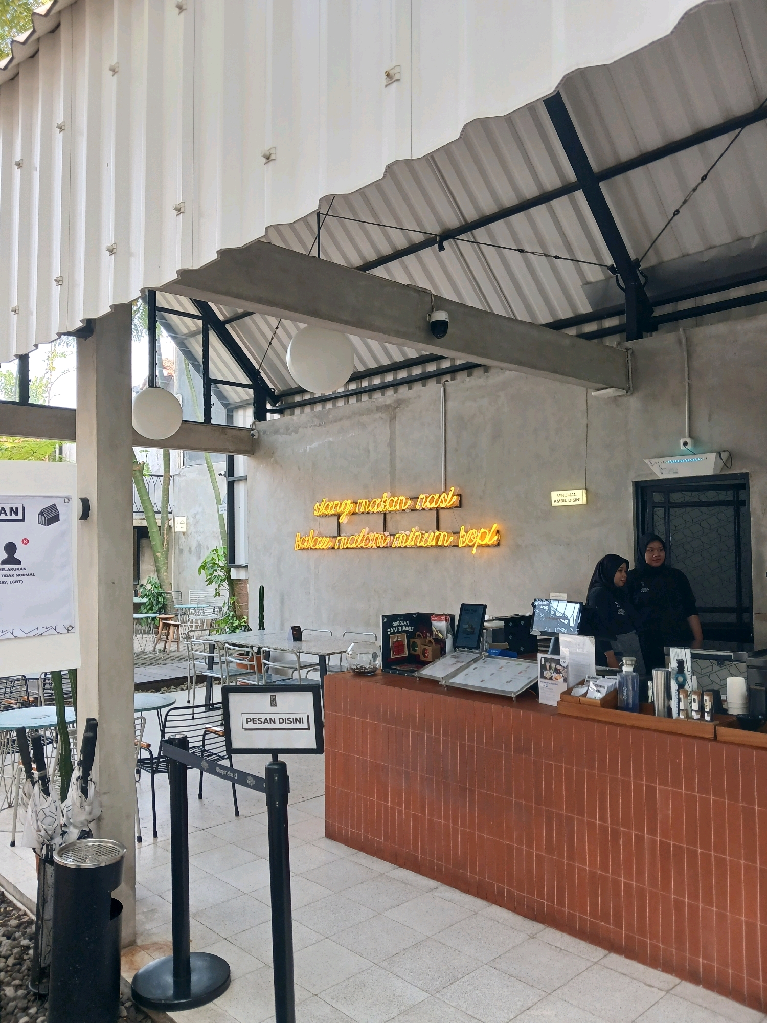 The image shows an outdoor cafe area. In the foreground, there is a counter with two women in black uniforms, presumably staff, standing behind it. On the counter, there are laptops, menus, and various bottles and containers. A neon sign on the wall behind the counter displays text in yellow. To the left of the counter, a stanchion with a "PESAN DISINI" (Order Here) sign is visible, along with an umbrella stand containing several umbrellas and a trash can. The cafe has a corrugated metal roof supported by concrete pillars. In the background, there are tables and chairs, suggesting a seating area. Trees and other buildings are visible further in the background. The overall scene depicts a modern, casual cafe environment.