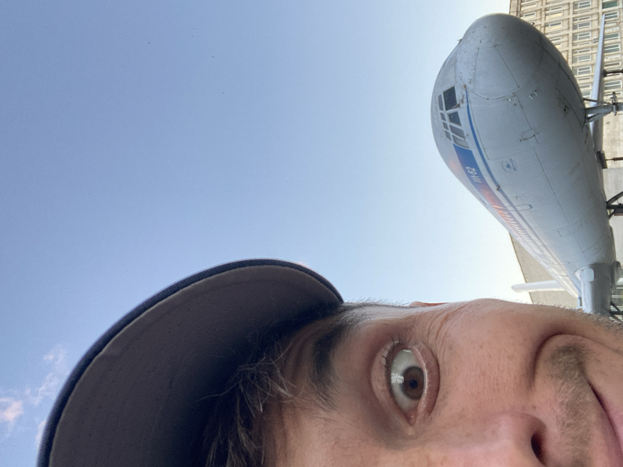 This is a first-person, low-angle, upside-down selfie taken outdoors during the daytime. The sky is a clear, light blue with a few wisps of white clouds. In the foreground, the edge of a dark baseball cap is visible, obscuring the top of the head. The majority of the image is dominated by the face of a person, captured in an extreme close-up. Their right eye, with a brown iris and visible reflections, is wide open and looking directly at the camera, conveying a sense of surprise or excitement. Their skin appears fair, with some visible pores and fine lines. They have dark eyebrows and some stubble on their face.
In the background, a large, grey airplane is prominently featured. The nose of the airplane, with a section of windows, is the most visible part. There is a blue stripe running along the side of the fuselage, and some white text appears to be written on it. The text "М-62" is clearly visible in blue lettering within the stripe. The airplane appears to be parked and possibly on display, as it is positioned in front of a tall, multi-story building with many windows. The building has a Brutalist architectural style. The overall scene suggests the person is visiting an aviation museum or an outdoor exhibition of aircraft, likely in Ulyanovsk, Russia, given the context provided. The lighting indicates it is a sunny day.