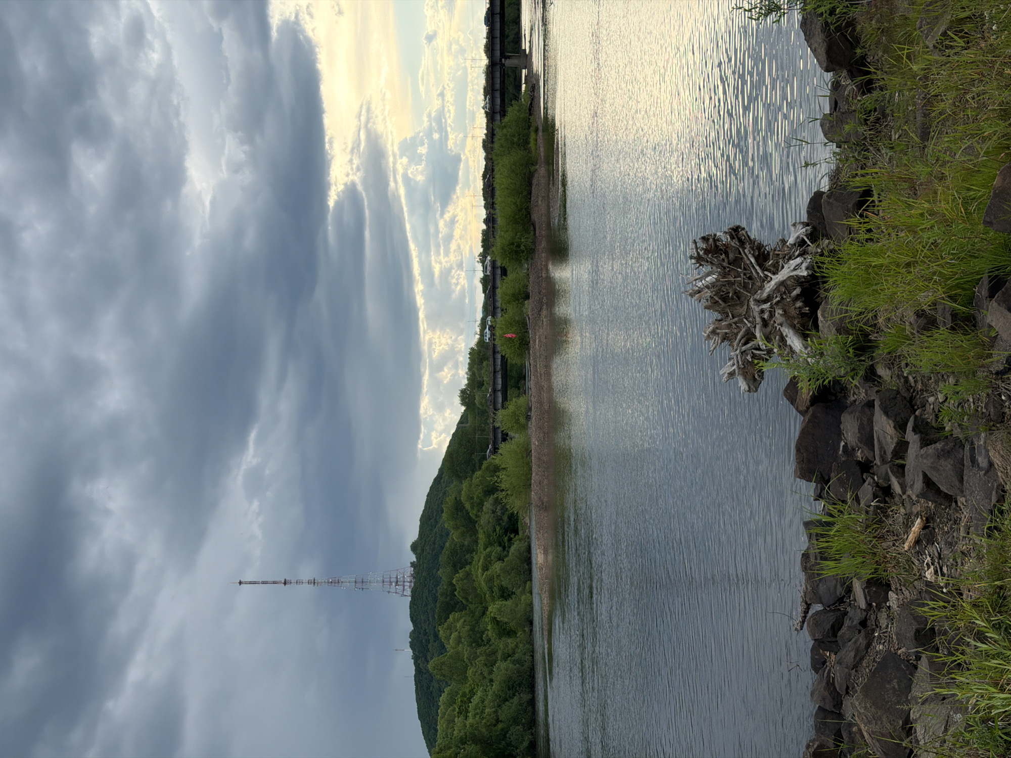 This landscape image captures a wide river or body of water on the right, bordered by a rocky and grassy bank. A large piece of light-colored driftwood or roots is visible on the bank near the water's edge in the foreground. The water's surface is rippled, reflecting the bright, cloudy sky above.
On the left side of the frame, a densely forested hill or mountain rises, covered in lush green trees. A tall, slender lattice transmission tower, red and white in color, stands prominently on the peak of this hill. In the middle ground, spanning across the water and above the tree line, a bridge structure is visible, likely carrying a road or railway, with what appears to be faint shapes of vehicles on it.
The sky above is dramatic, with heavy, dark grey clouds dominating the upper left portion, while the right side of the sky shows brighter, partially illuminated clouds, suggesting the sun is breaking through. The lighting indicates late afternoon or early evening, casting a soft, slightly golden glow on the water and the brighter cloud formations. No specific activity or visible text is present in the scene. The overall atmosphere is tranquil and expansive.