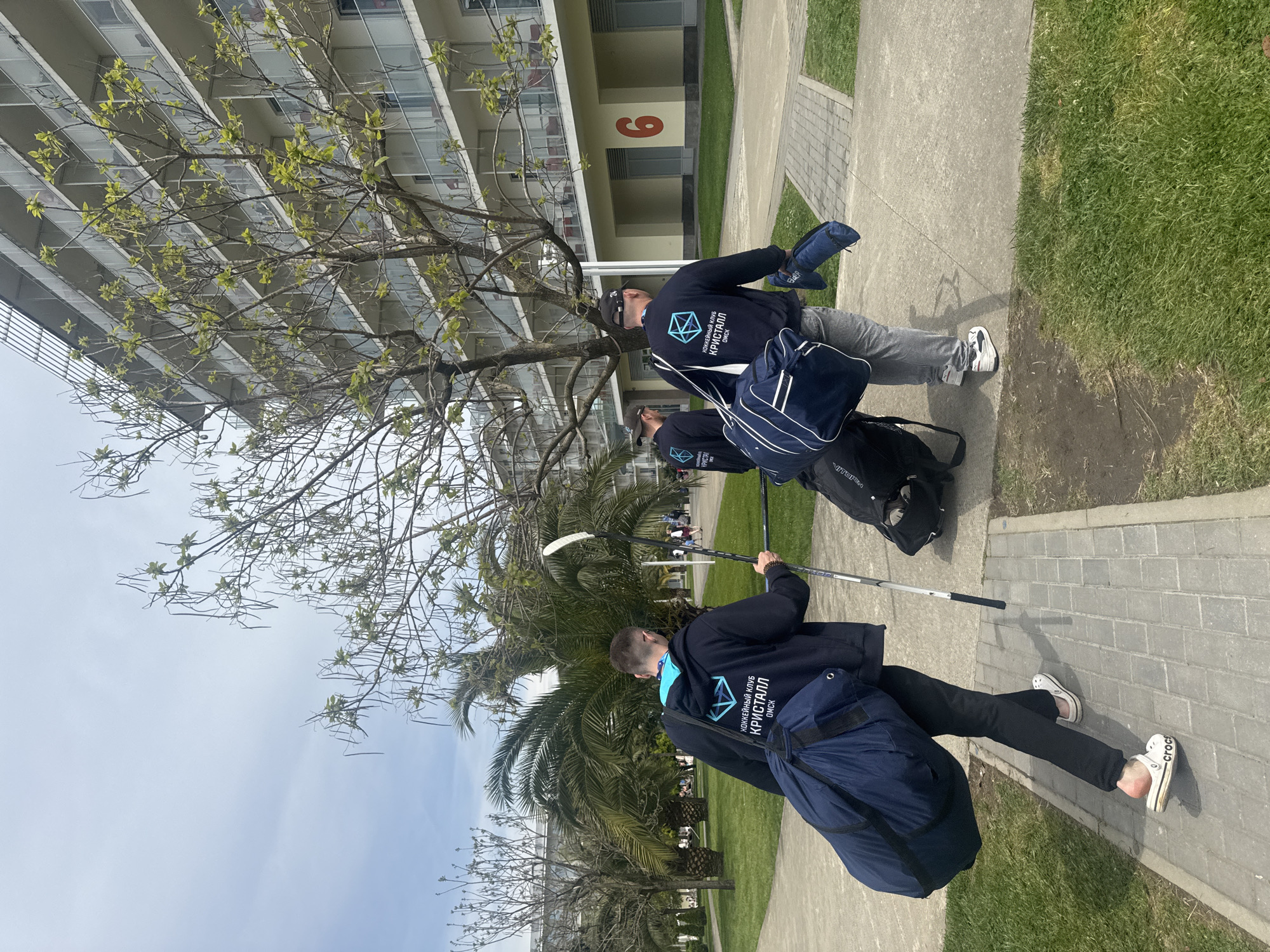 The scene depicts three individuals walking along a paved walkway alongside a grassy area. The location is Sirius, Russia, as evidenced by the building with the number "6" visible in the background. The individuals wear dark jackets with the logo of "КРИСТАЛЛ Oмск" (Kristall Omsk), suggesting a sports team affiliation. Each person carries a large navy blue bag and appears to be holding hockey sticks. The setting is outdoors on a sunny day, with buildings and trees visible in the background. The individuals are walking, and it seems they are associated with a sports team, likely ice hockey.