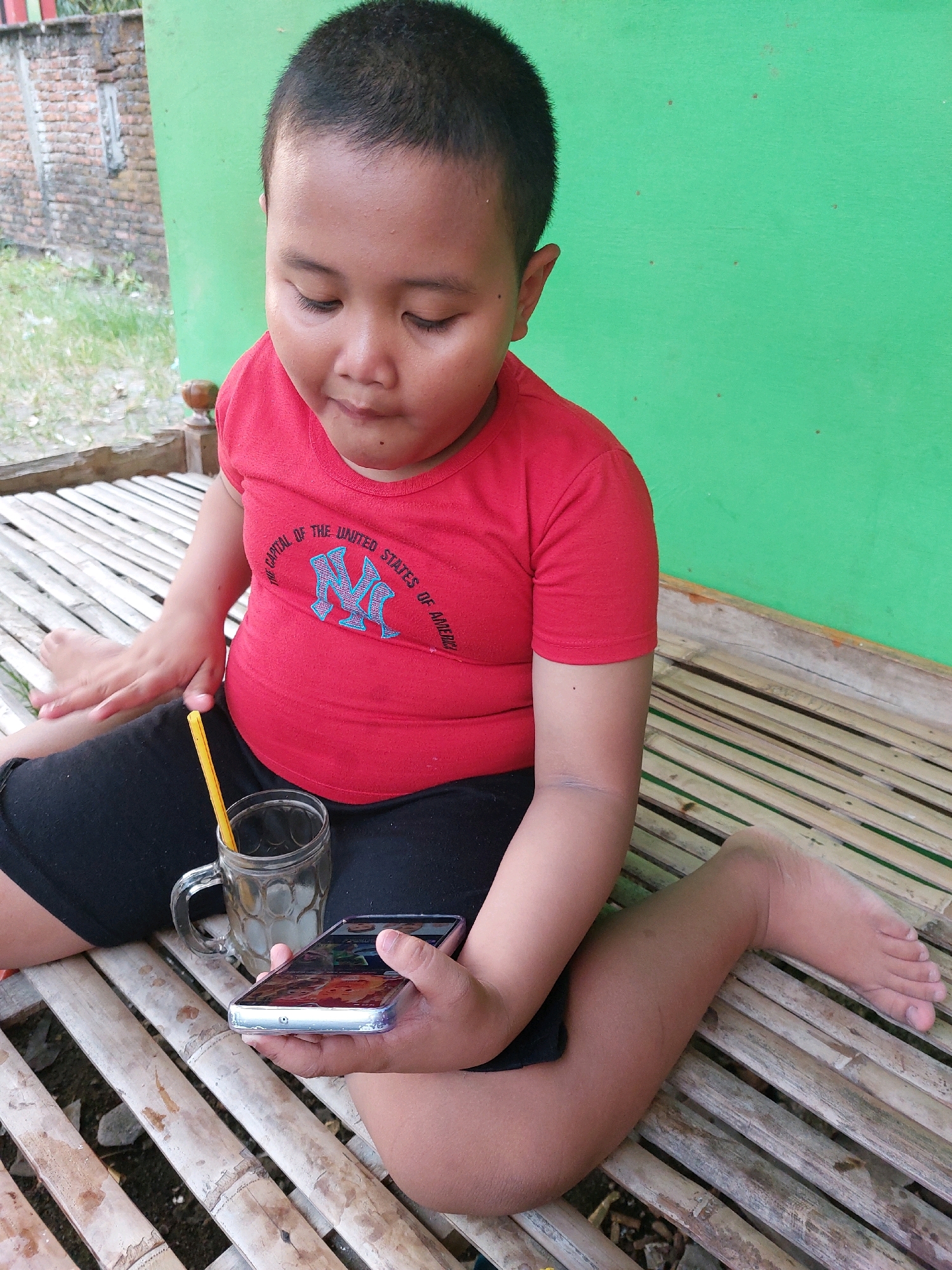A young boy, wearing a red t-shirt with a New York Yankees logo and black shorts, sits on a bamboo bench outdoors. He is holding a smartphone in his right hand, looking down at the screen. A glass of a light-colored beverage with a yellow straw sits next to him on the bench. In the background, a green wall and a brick structure are visible, suggesting an outdoor setting in a residential or public area. The boy's posture and gaze indicate he is engaged with the device.