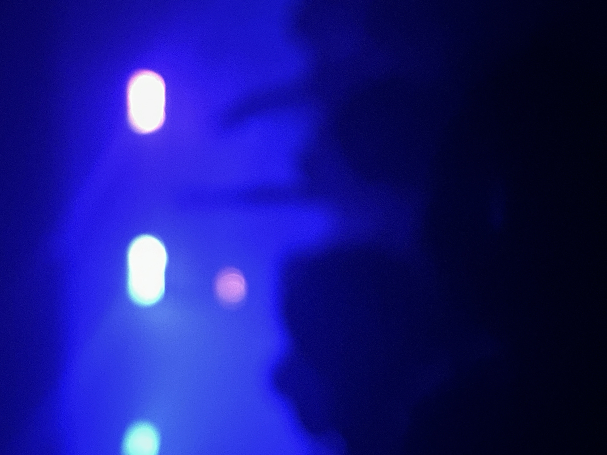 The image shows a blurred, low-light scene with blue and dark blue hues dominating the color palette. There are three prominent out-of-focus light sources appearing as bright, blurry ovals. In the right half of the image, there are indistinct dark shapes, possibly suggesting shadows or obscured objects/figures. It is not possible to definitively determine the location in Paris, France, or any specific details about people, subjects, objects, actions, or interactions due to the extreme blur and lack of clarity. The environment appears to be dark, possibly indoors, based on the lighting and lack of recognizable features.