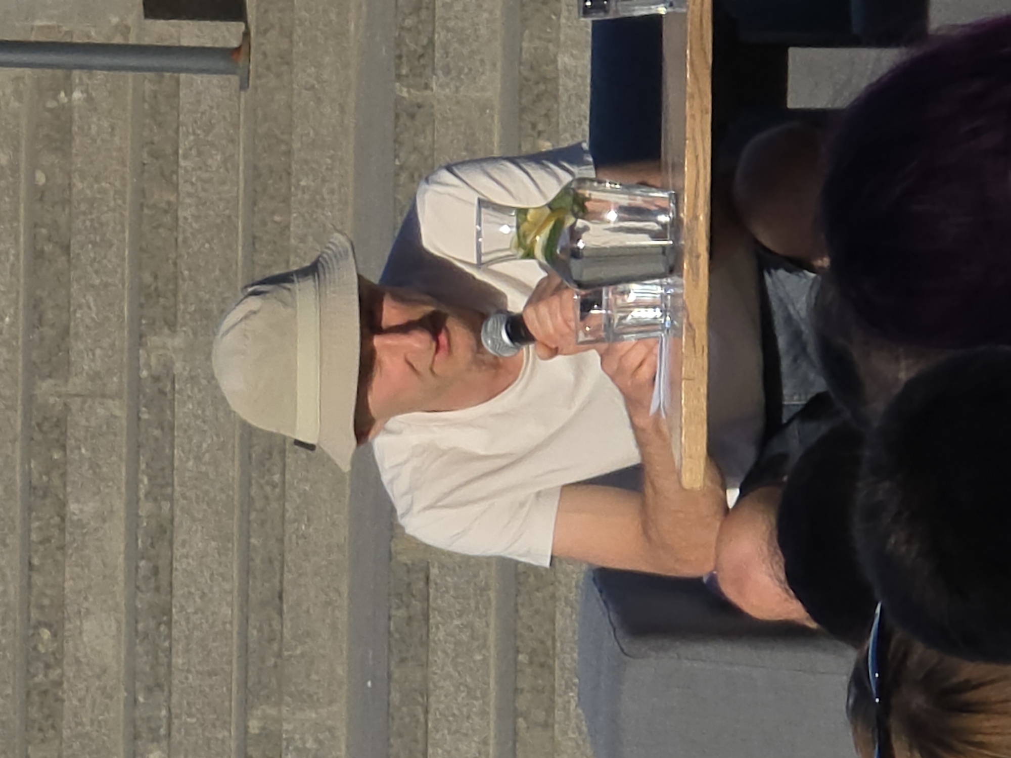 A medium-shot photo depicts a middle-aged Caucasian man speaking into a microphone while standing behind a table. He is wearing a white t-shirt and a tan bucket hat. A glass bottle filled with water and slices of lime or lemon sits on the table. In the background are stone steps. To the right are heads of other people. The photo was taken at Warsaw, Poland.