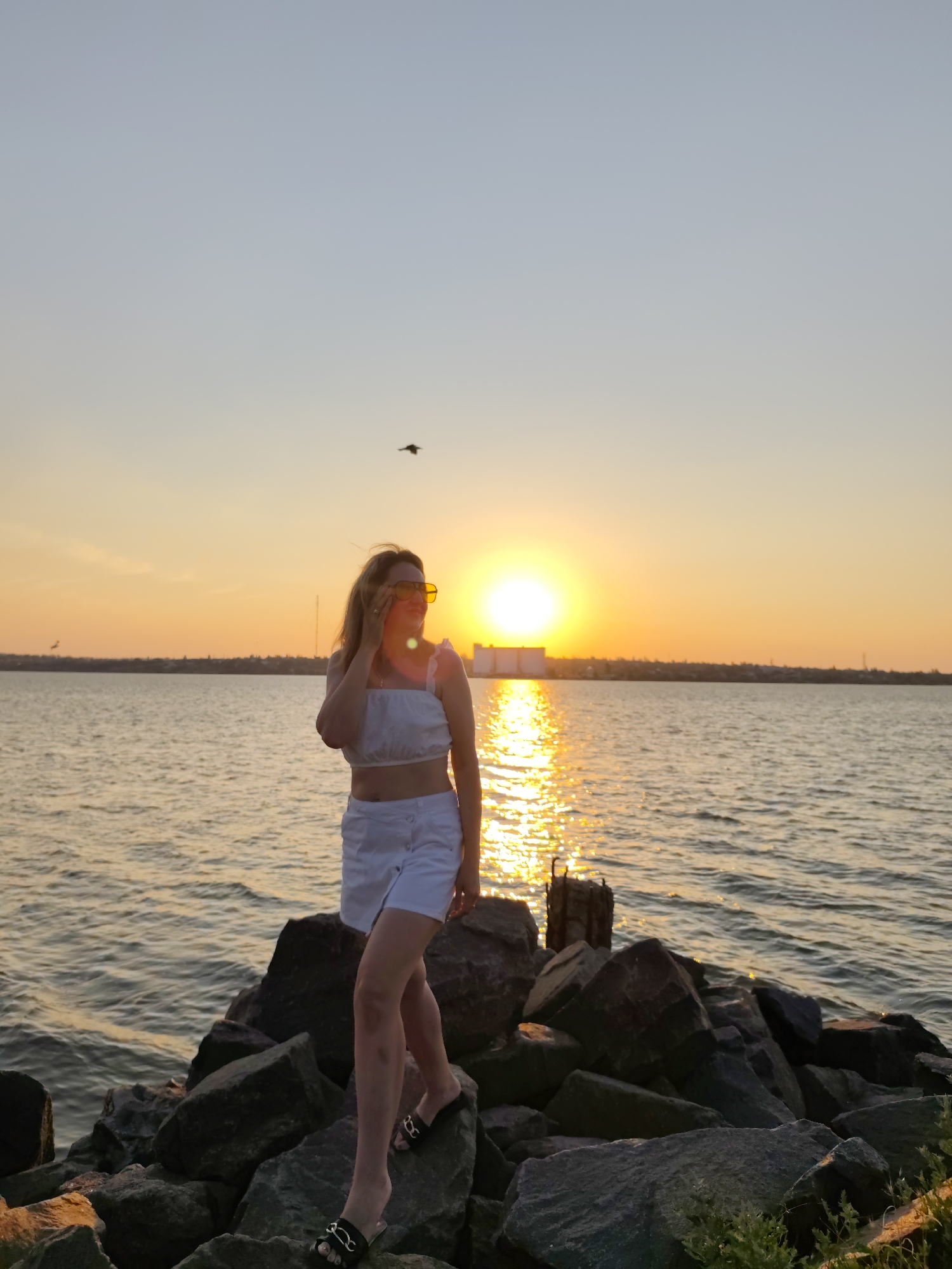 This media file captures a woman standing on a rocky shore during sunset. She is wearing a white crop top and white shorts, with black slip-on sandals. Her hair is blonde, and she is wearing yellow sunglasses, with one hand near her face. The woman appears to be posing for the photograph, looking towards the right side of the frame.
The setting is a body of water, likely a sea or large river, with a distant shoreline visible. The sky is filled with the warm hues of sunset, with the sun itself low on the horizon, casting a golden reflection on the water. A building or industrial structure is visible on the far shore, silhouetted against the bright sky. There is a single bird flying in the sky above the woman.
The overall mood of the image is serene and tranquil, enhanced by the beautiful sunset. The rocky shore provides a rugged contrast to the calm water and the peaceful atmosphere. The time of day is clearly sunset, indicated by the low sun and the colors in the sky. The location is indicated as Mykolaiv, Ukraine. There is no visible text in the media file.