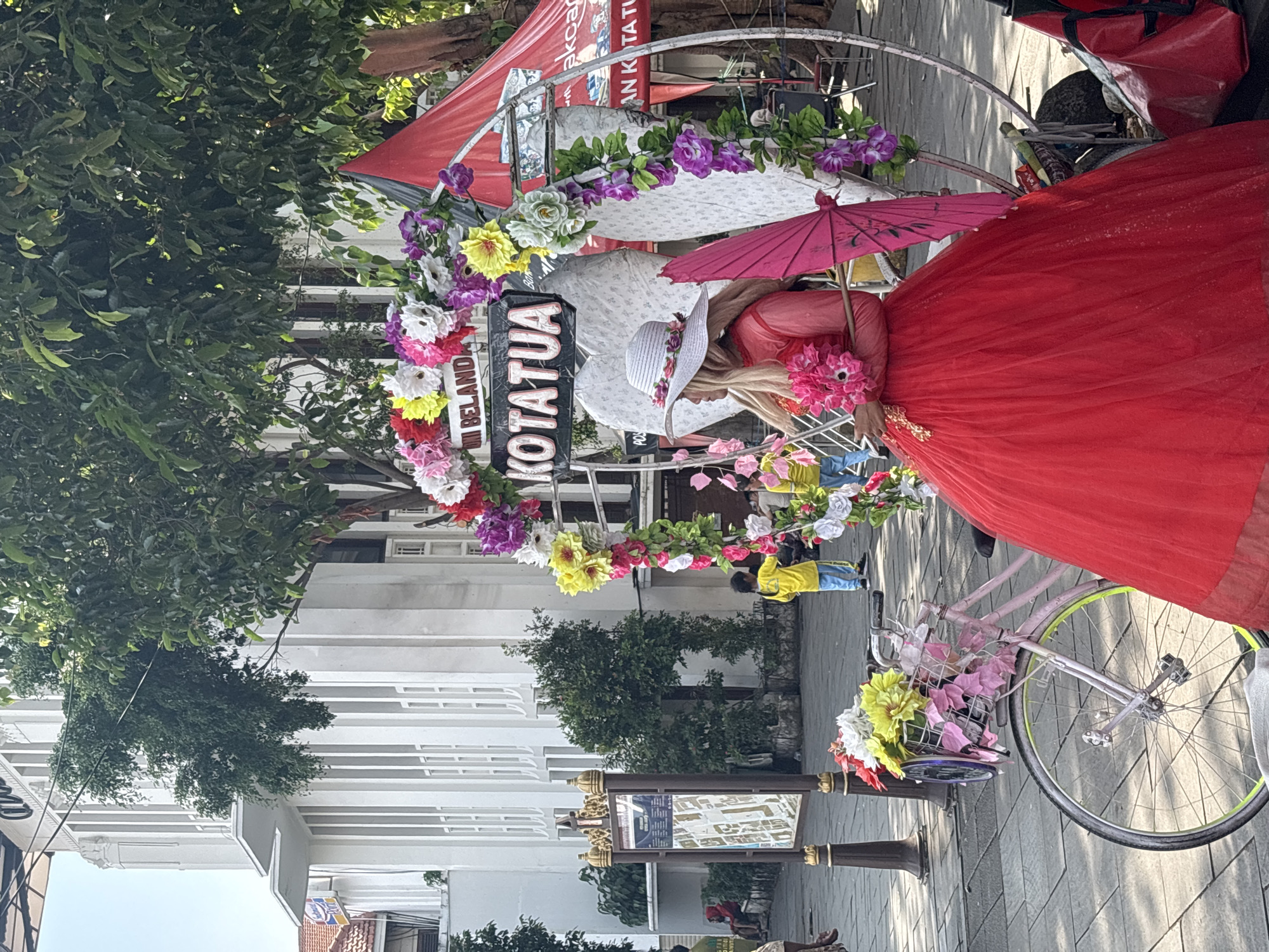 A person in a long red dress and wide-brimmed hat stands next to a decorated bicycle rickshaw in Kotatua, West Jakarta, Indonesia. The rickshaw is adorned with flowers and a sign that says "KOTATUA." The scene includes a street setting with buildings, trees, and other people in the background.