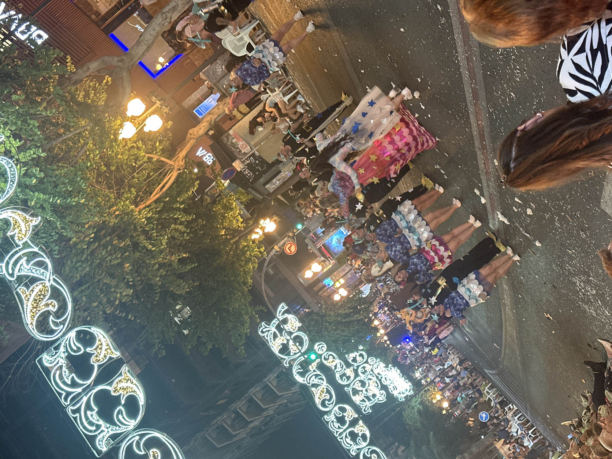 The image depicts a nighttime street scene, likely a parade or festival, in Carrer de Castanyos (Castanos Street) in Alicante, Spain. The perspective is slightly elevated and angled, looking down the street.
In the foreground, the backs of two spectators' heads are visible, partially obscuring the bottom right. The street surface, covered in small white confetti pieces, stretches into the midground. A procession of individuals in various costumes is advancing along the street, moving from right to left across the frame. Many participants wear coordinated outfits featuring blue long-sleeved tops and short, ruffled skirts, some white-silver and others bright pink or fuchsia. One person wears a flowing white dress adorned with blue stars. Another individual is dressed in a dark, possibly black, full-body costume with yellow accents and a mask, holding a staff-like object.
Along the left side of the street, spectators are seated on rows of white plastic chairs, observing the parade. In the background, multi-story urban buildings line the street. A prominent illuminated "BBVA" sign is visible on a building to the right, along with other partially visible commercial signs. Streetlights with multiple warm-toned bulbs provide illumination, complemented by elaborate, ornate white and gold illuminated decorative light structures suspended overhead across the street. Green foliage from trees is interspersed along the street. Further down the road, a green traffic light and a red circular street sign with an arrow are identifiable. The entire visible road surface is heavily strewn with confetti or similar celebratory debris.
