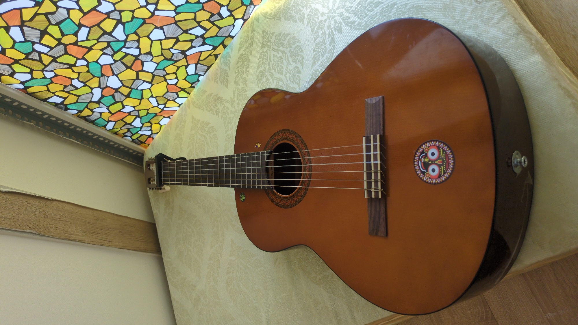 A brown classical guitar lies on a yellow patterned surface, likely a table or cushion. The guitar has six strings and a sticker of two eyes surrounded by a colorful patterned circle. Behind the guitar is a stained-glass-like window in shades of yellow, orange, green, white, and black. The surroundings appear to be an interior space. The scene doesn't allow for a specific identification of Da Lat, Vietnam.