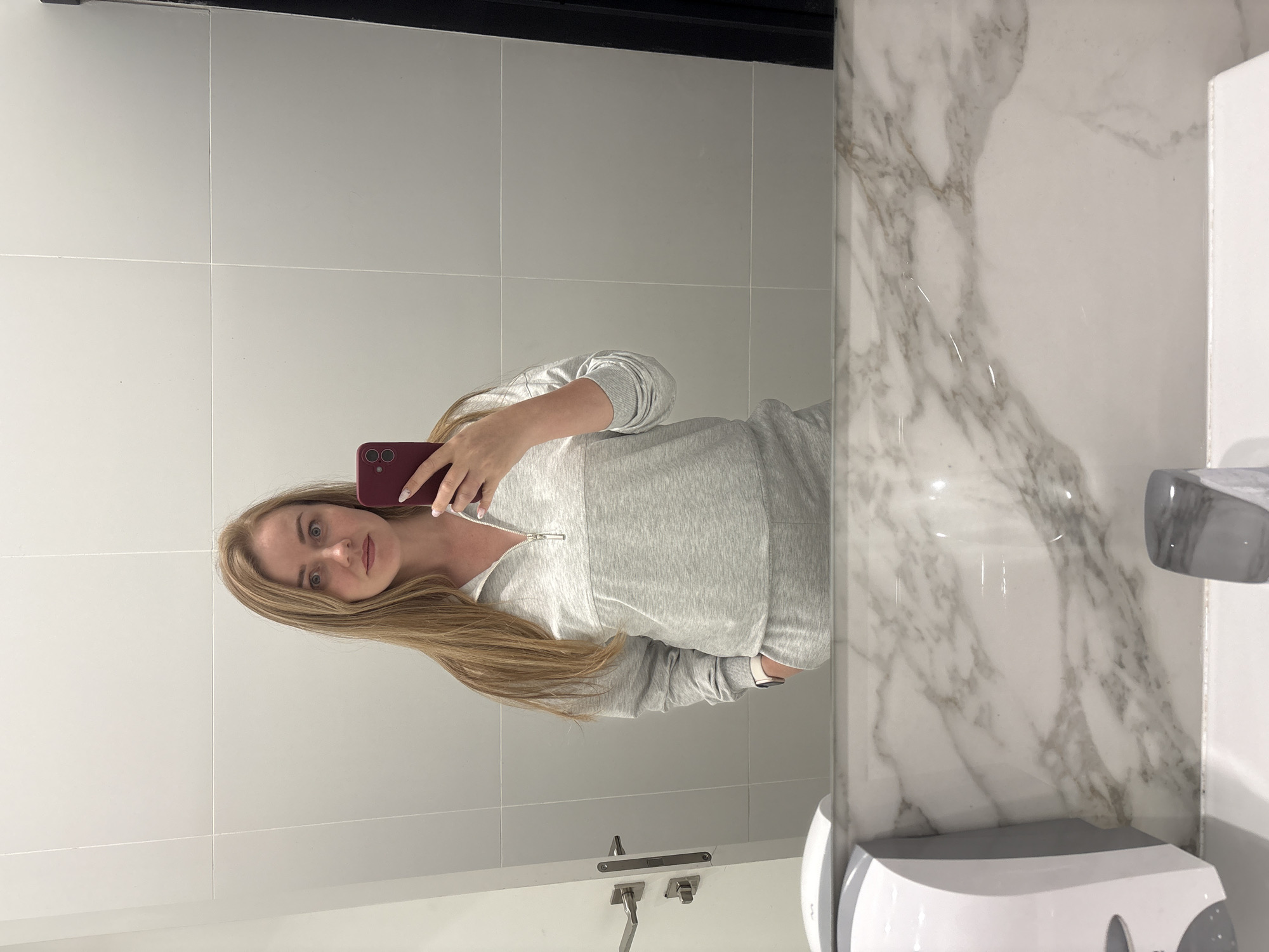 A blonde woman takes a selfie in a bathroom mirror. She is wearing a grey sweatshirt and holding a red phone. The bathroom has white tile walls, a marble countertop, and a white hand dryer. There's a tap and handwash facility is visible as well. No additional information about the location of Новое Мартьяново, Russia, is discernible from the image.