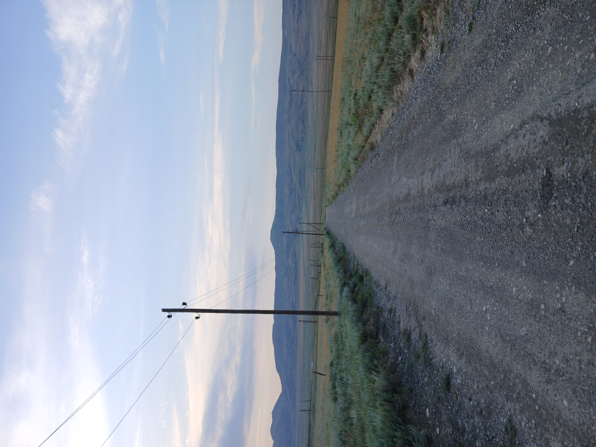 An overhead shot shows a dirt road stretching into the distance, flanked by grassy fields and rolling hills under a pale blue sky. A telephone pole stands tall beside the road, its wires disappearing into the distance. The scene suggests a rural setting in Yanasha, Russia.