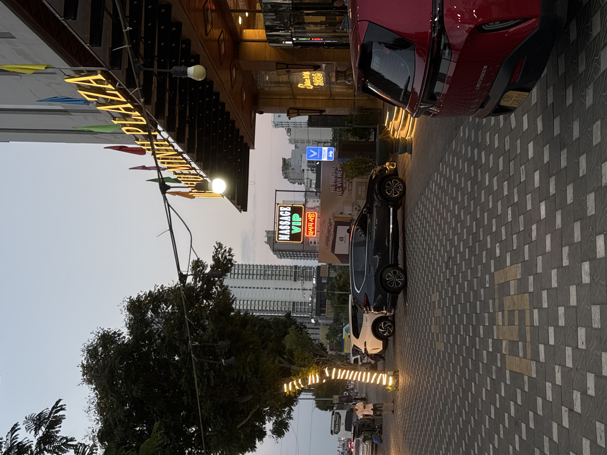 The image depicts the exterior of the "DIAMOND PLAZA" building in Đà Nẵng, Vietnam, at dusk. The building's facade features a prominent illuminated sign that spells out "DIAMOND PLAZA" in large yellow letters. Several colorful flags (yellow, blue, green, red) are attached to the building above the sign. A neon sign for "MASSAGE VIP" is also visible in the background.
In the foreground, a paved walkway with a patterned brick surface is present. A dark gray SUV and a white SUV are parked on the right side of the image, facing away from the viewer. A red car, with its rear end visible on the far right, is also parked. A street lamp decorated with white lights illuminates a tree on the left side of the image. A person in a light-colored shirt and shorts is standing near the tree, looking towards the street. A bus with "Vina Travel" written on it is visible in the distance on the left. The sky is a pale, clear dusk. The general environment appears to be an urban setting with commercial buildings and a street.