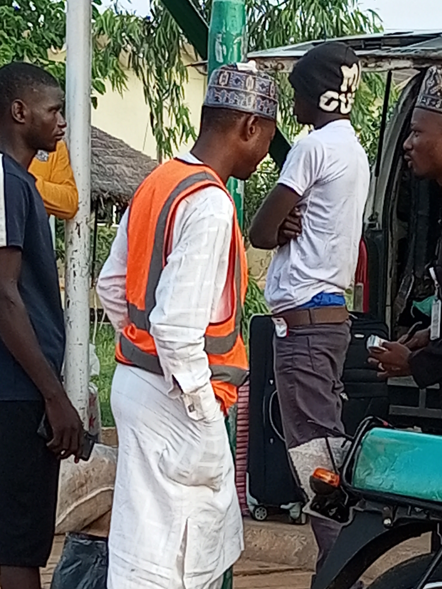 The image shows a group of men gathered outdoors, with a truck or van visible on the right. The man in the foreground, wearing a white robe and an orange safety vest, is standing with his hands in his pockets. To his left, another man in a blue shirt is looking away from the camera. On the right, a man in a white t-shirt and dark pants is standing with his arms crossed, and behind him, another man is partially visible. Luggage and a motorcycle are also present in the scene. The environment suggests an urban or semi-urban setting, possibly a transport hub or a roadside stop.