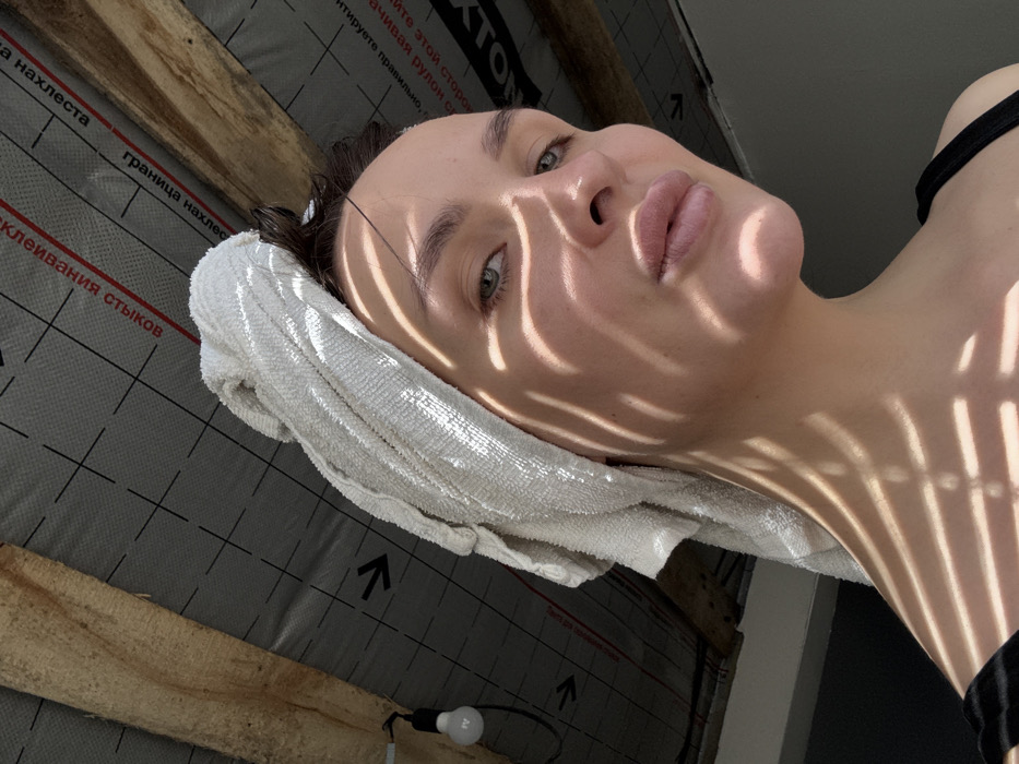 A female individual is depicted in a close-up, head-and-shoulders portrait. She has fair skin, light-colored eyes, and light pink lips. Her dark hair is wrapped in a white terry cloth towel. A black strap, possibly from a tank top, is visible on her right shoulder. Her face, neck, and the towel are marked by distinct, parallel striped shadows, consistent with light passing through slatted blinds or a similar obstruction. The background reveals an unfinished interior space, likely an attic or construction area. Exposed wooden beams are visible. Above these beams is a grey synthetic membrane or vapor barrier with a black grid pattern and red Russian text, including "КЛЕИВАНИЯ СТЫКОВ" (joint bonding) and "ГРАНИЦА НАХЛЕСТА" (overlap border). A single, bare incandescent light bulb hangs from a black cord in the upper left quadrant of the frame. The scene is located in Chaikovsky, Russia.