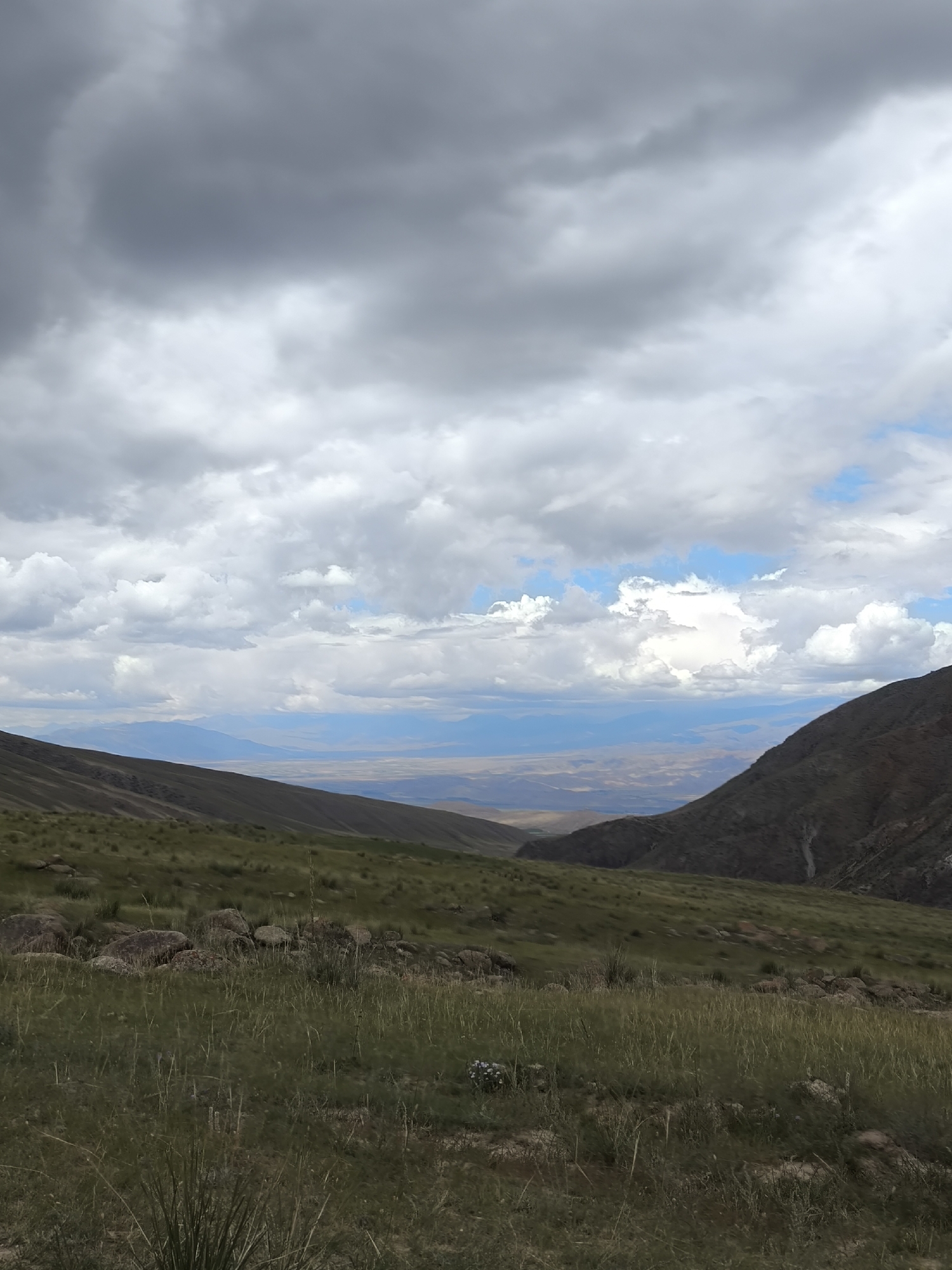 This is a vertical, eye-level, medium shot of a mountain valley in Bolshevik, Kyrgyzstan. The foreground is dominated by a grassy slope with scattered rocks. Two dark mountains frame the sides of the valley, with a hazy, arid landscape visible in the distance. The sky is filled with dramatic, gray and white clouds, with patches of blue peeking through. There are no people or animals visible in the image. The scene is natural and undeveloped, suggesting a remote or rural location.